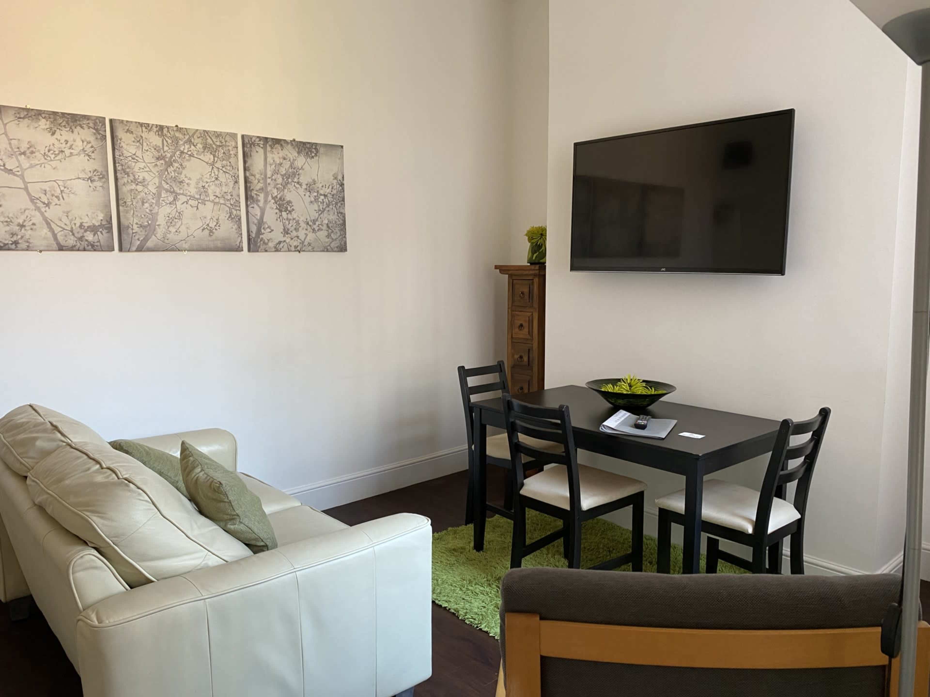 A small living area featuring a white couch, a black dining table with chairs, a television mounted on the wall, and three framed pictures.