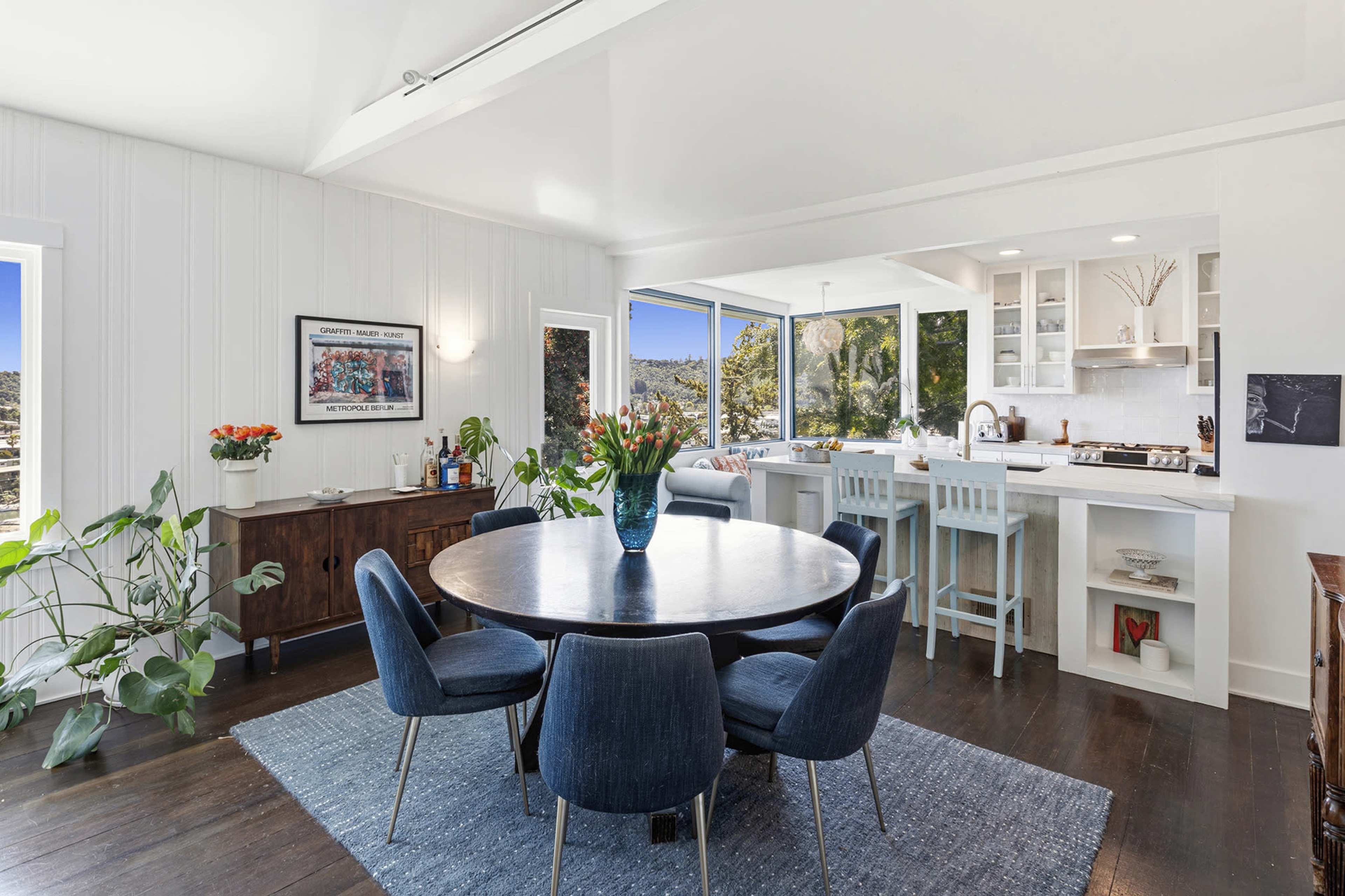 A modern dining area features a round table surrounded by upholstered chairs, with a view of large windows and an adjoining kitchen space.