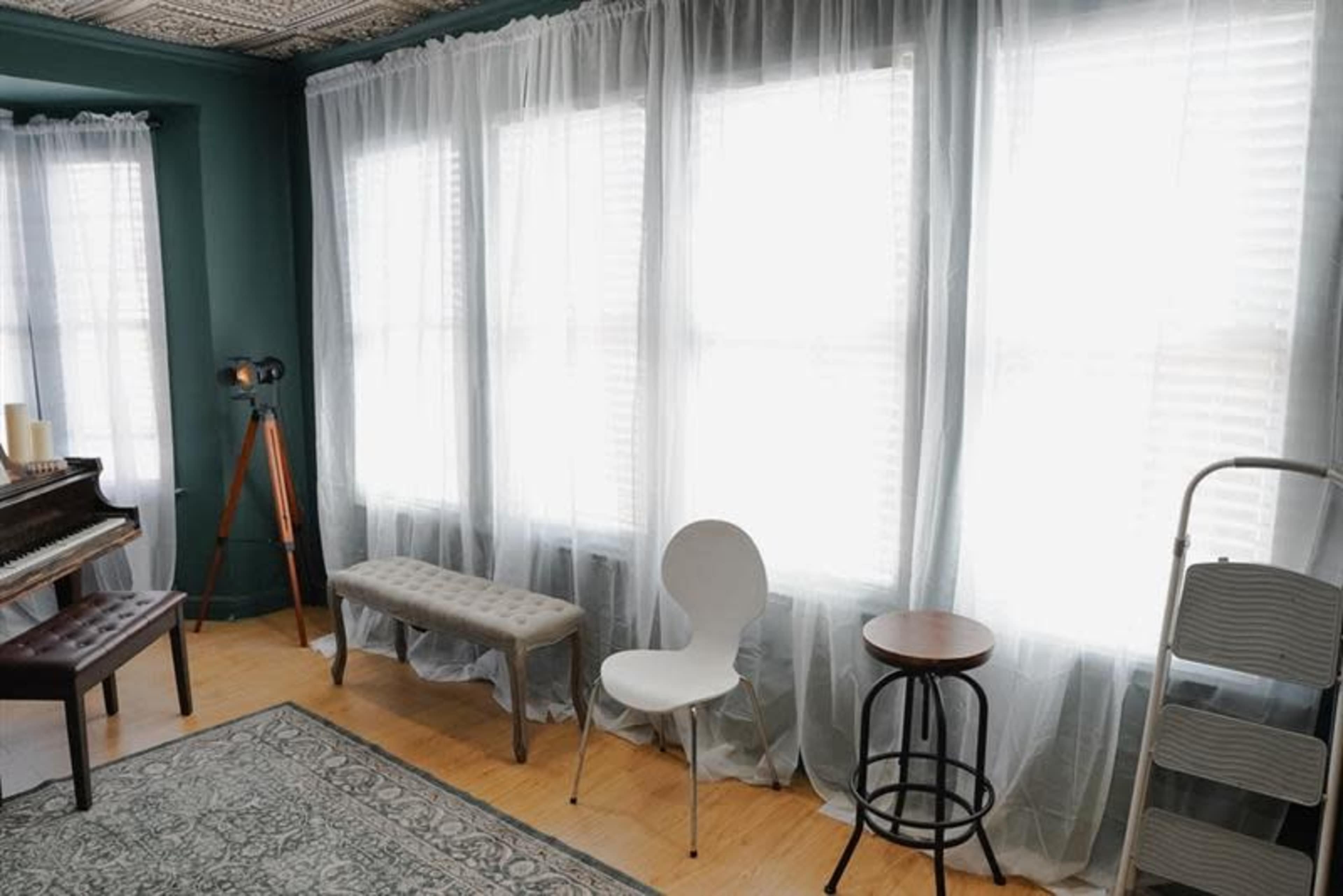 The image shows a room with large windows draped in sheer white curtains, featuring a bench, two chairs, a small round table, and a tripod standing lamp.