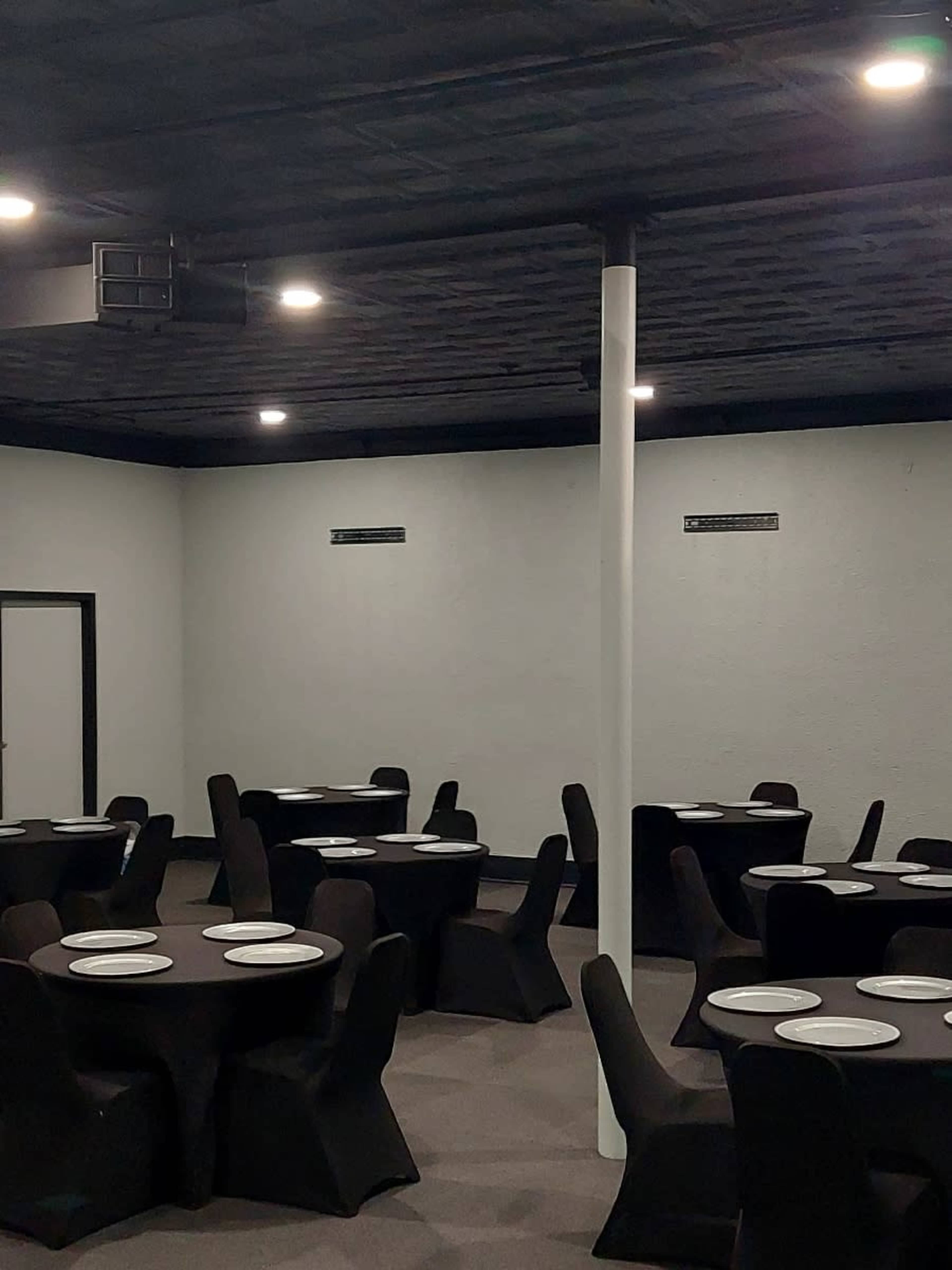 The image shows a dining area with several round tables covered by black tablecloths and white plates set on top.