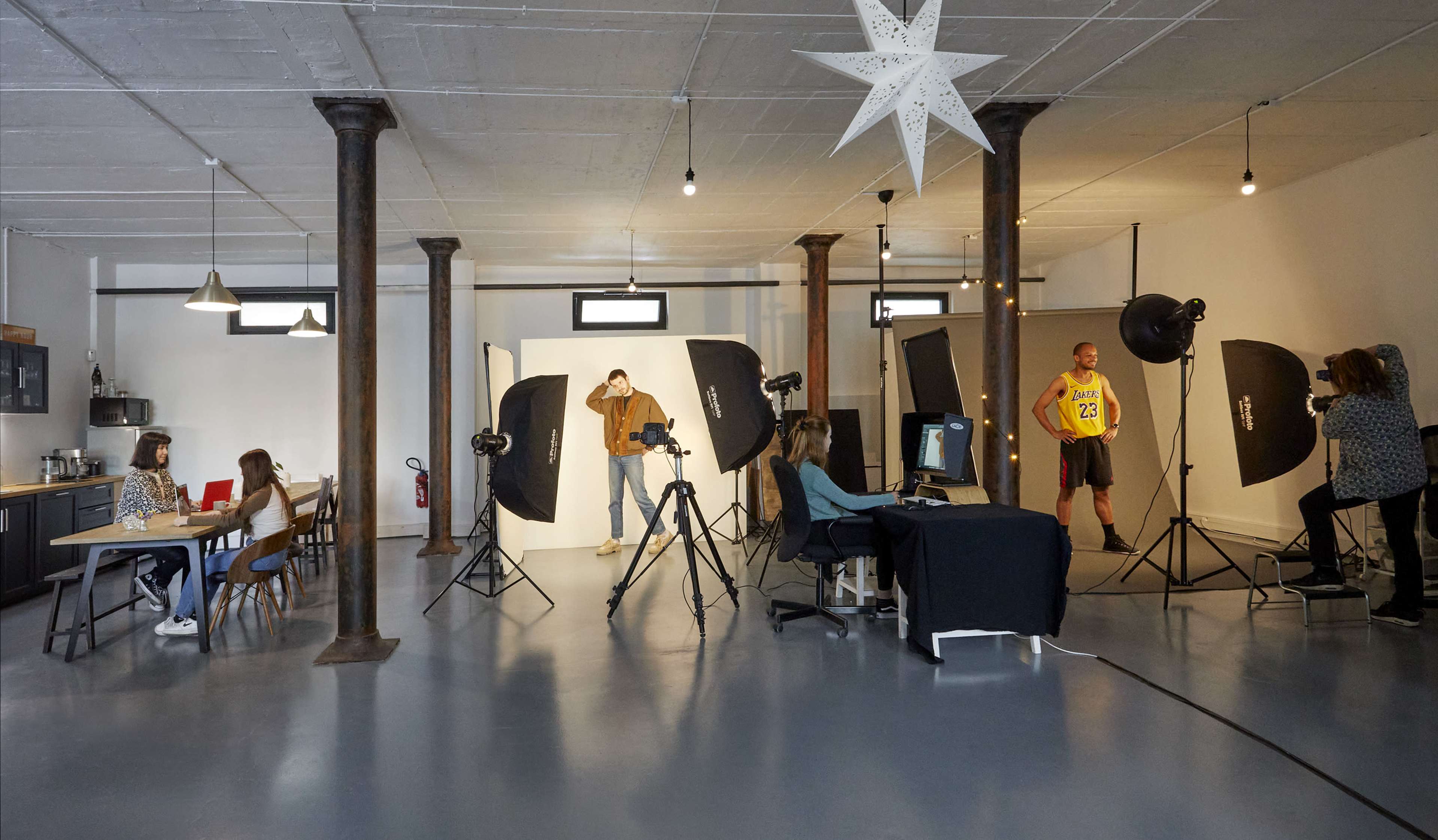 A photography studio features a model posing against a backdrop, cameras, lighting equipment, and a team of people working at tables and monitors.