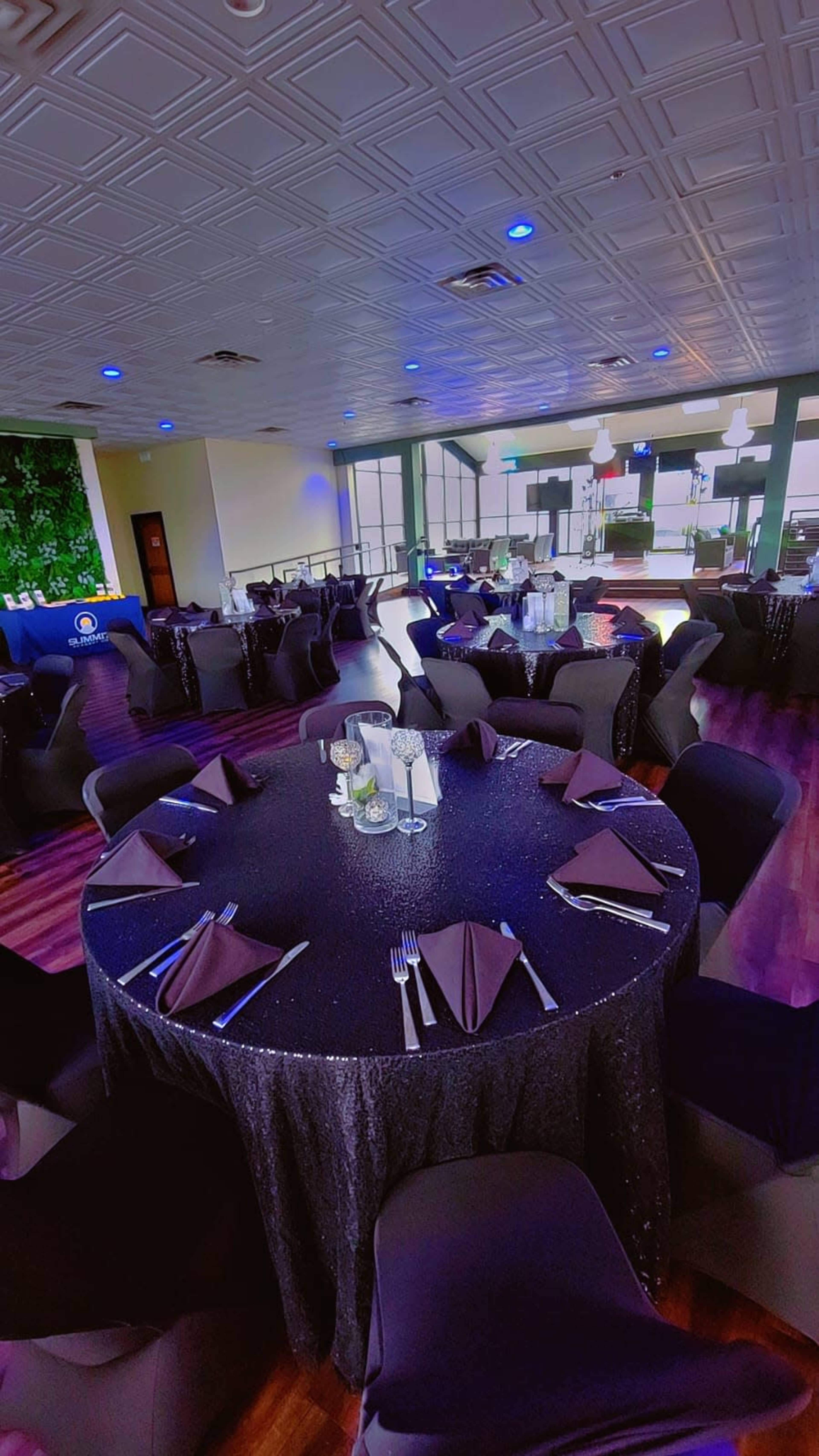 The image shows a modern dining space with several round tables covered in black tablecloths, set with utensils and napkins, in a room featuring green accents and colorful lighting.