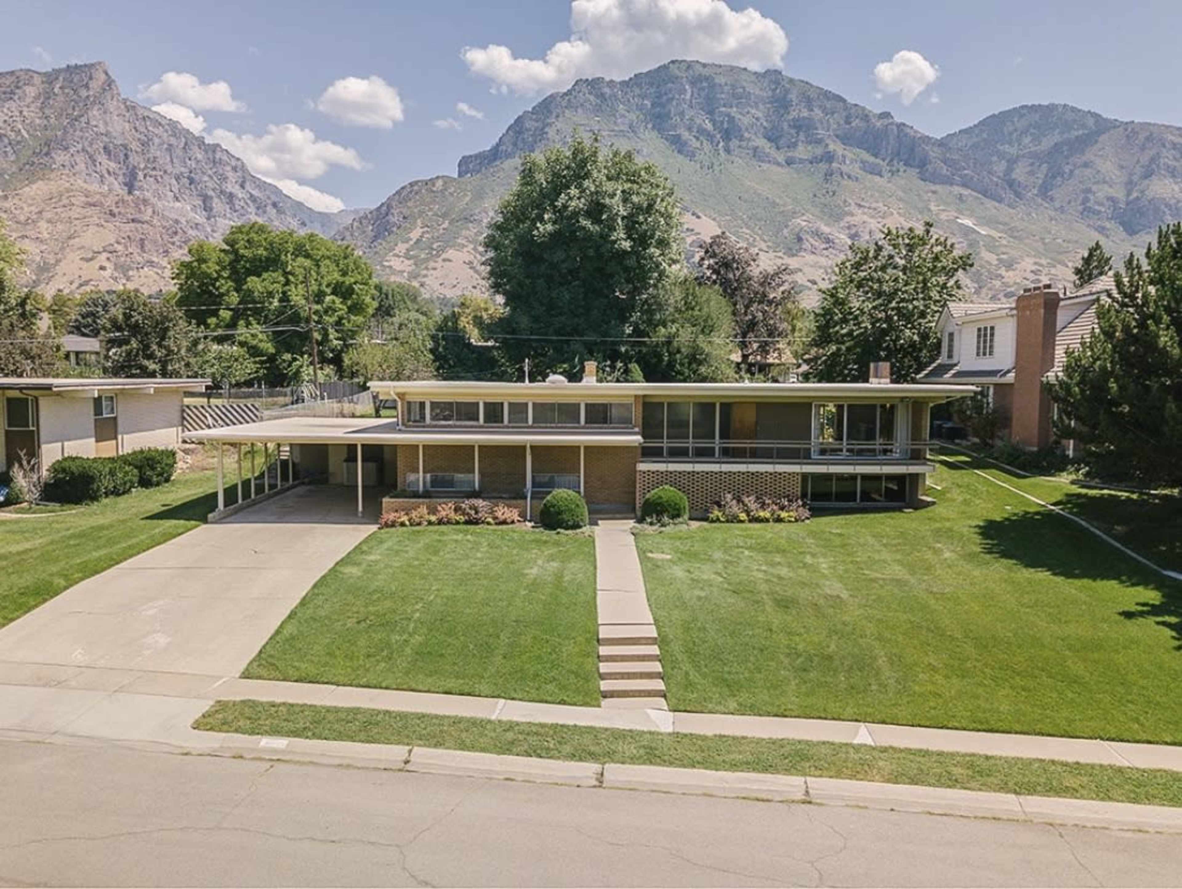 A modern, one-story house with a brick facade sits on a well-manicured lawn, framed by mountains in the background.