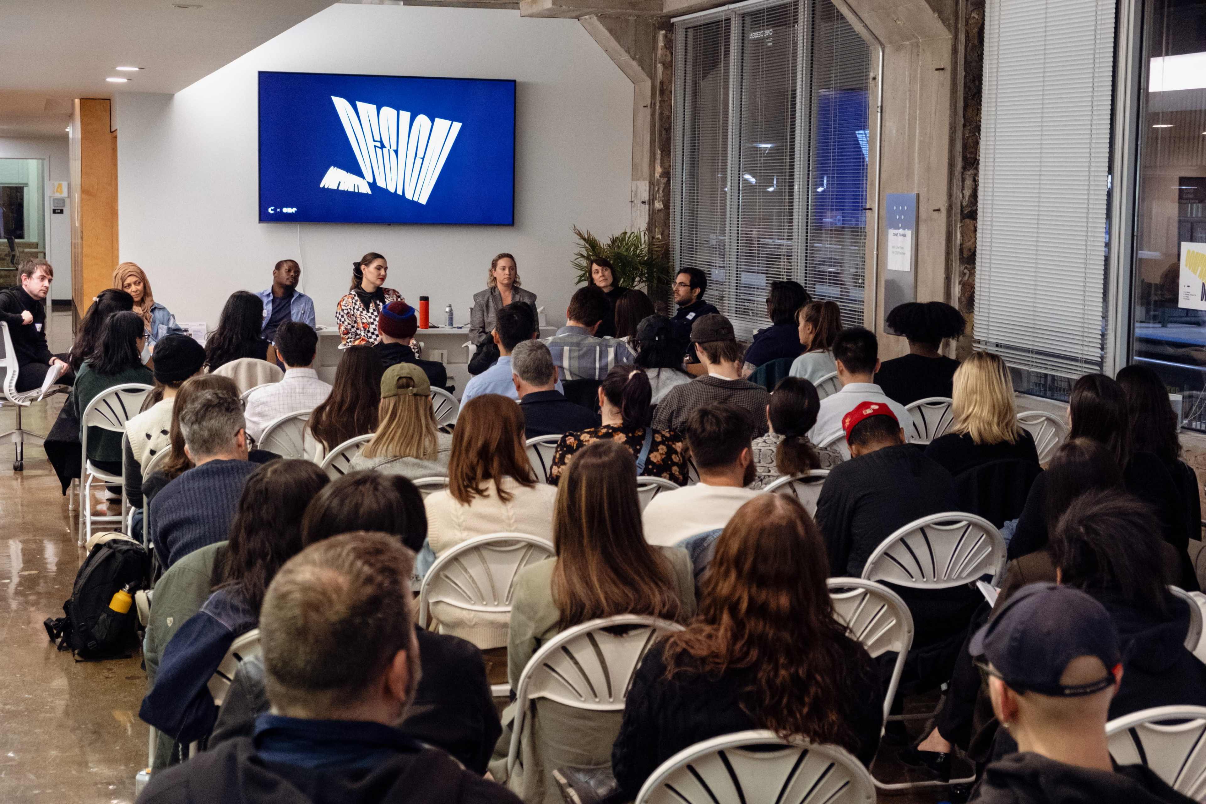 A group of people is seated in chairs facing a large screen displaying the word "DESIGN" during a panel discussion event.