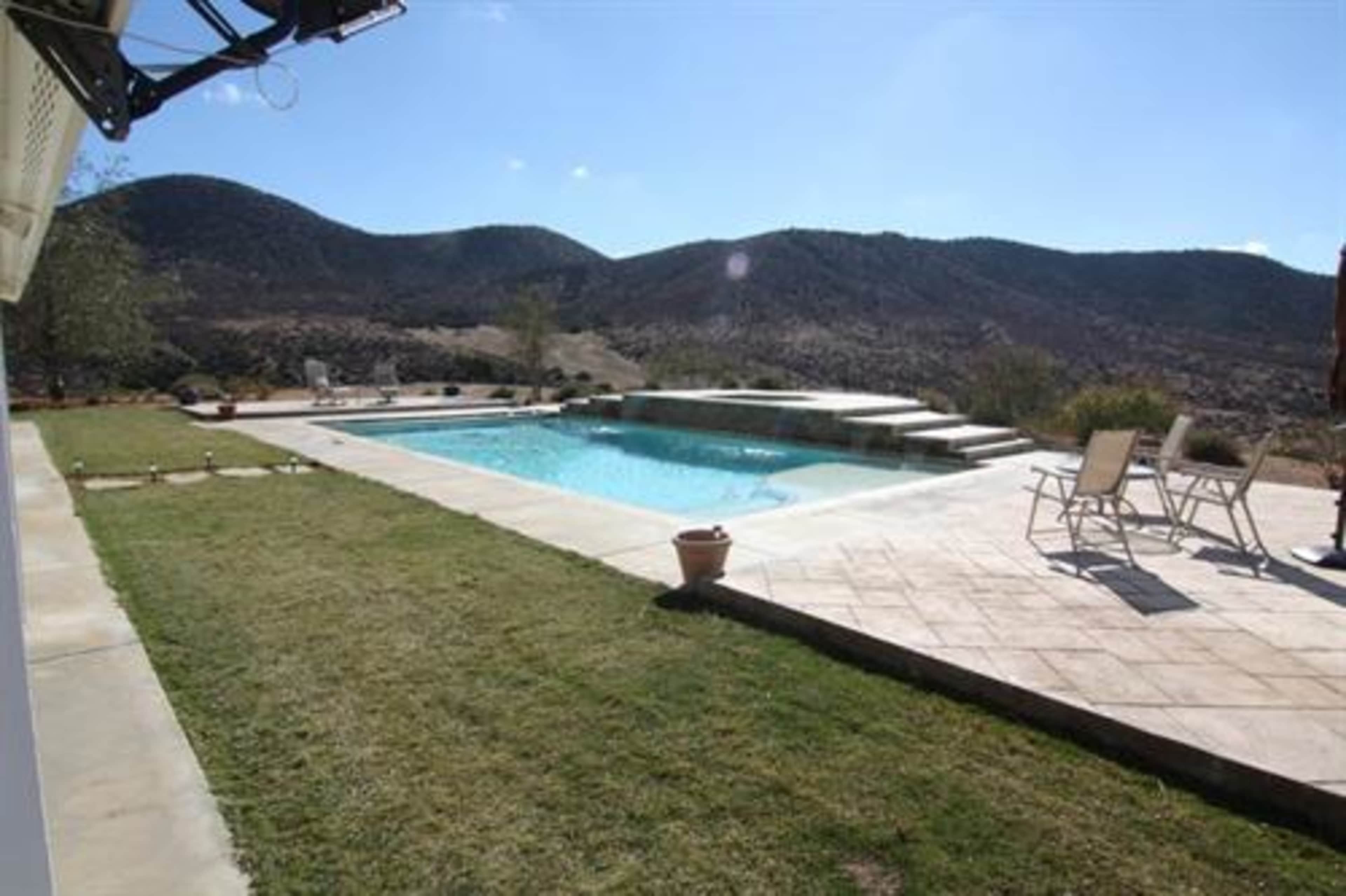 A swimming pool surrounded by a patio and grass, with mountains in the background under a clear sky.