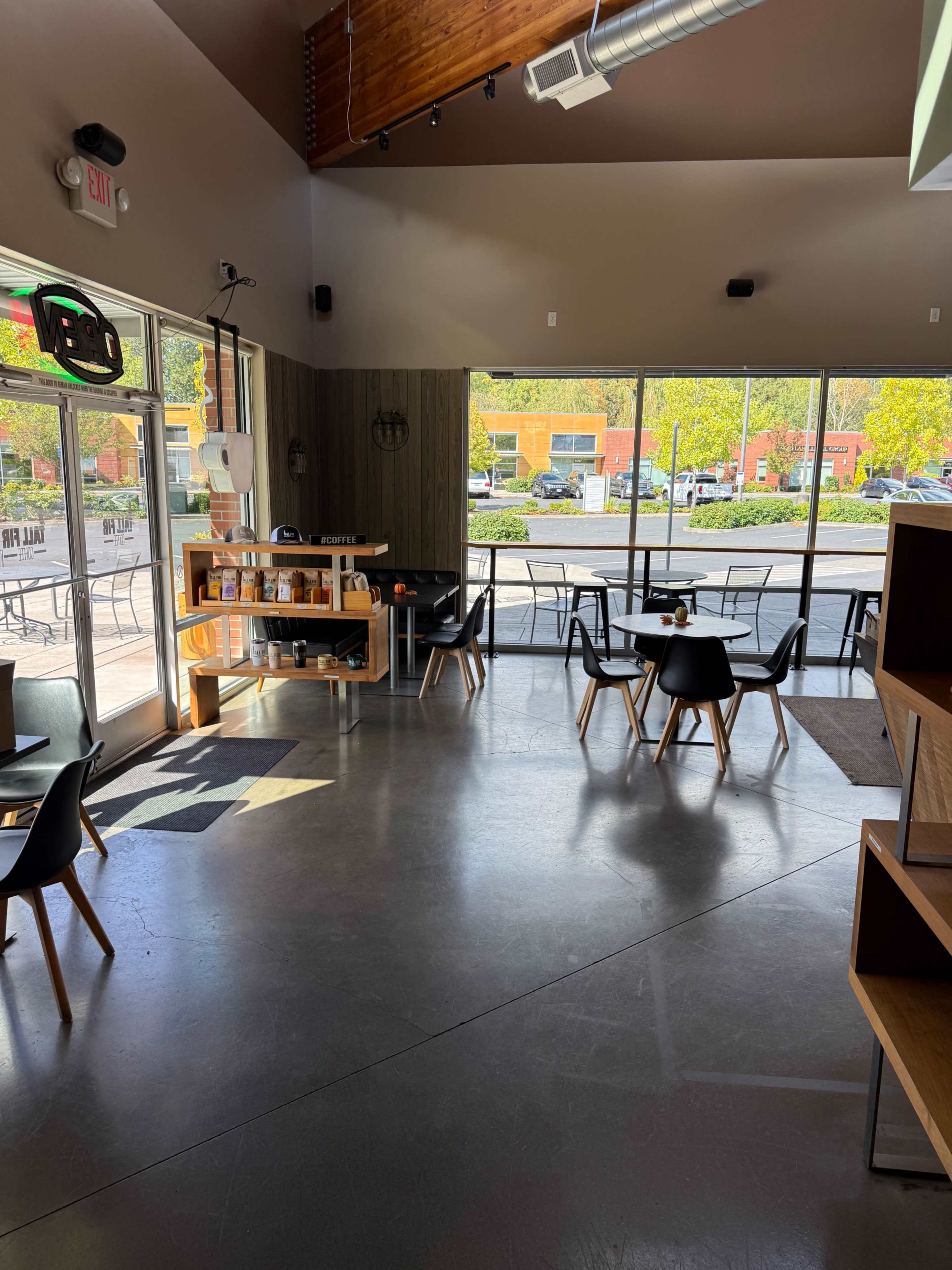 The image shows a modern cafe with a spacious interior featuring black chairs and tables, large windows allowing natural light, and a view of an outdoor seating area.