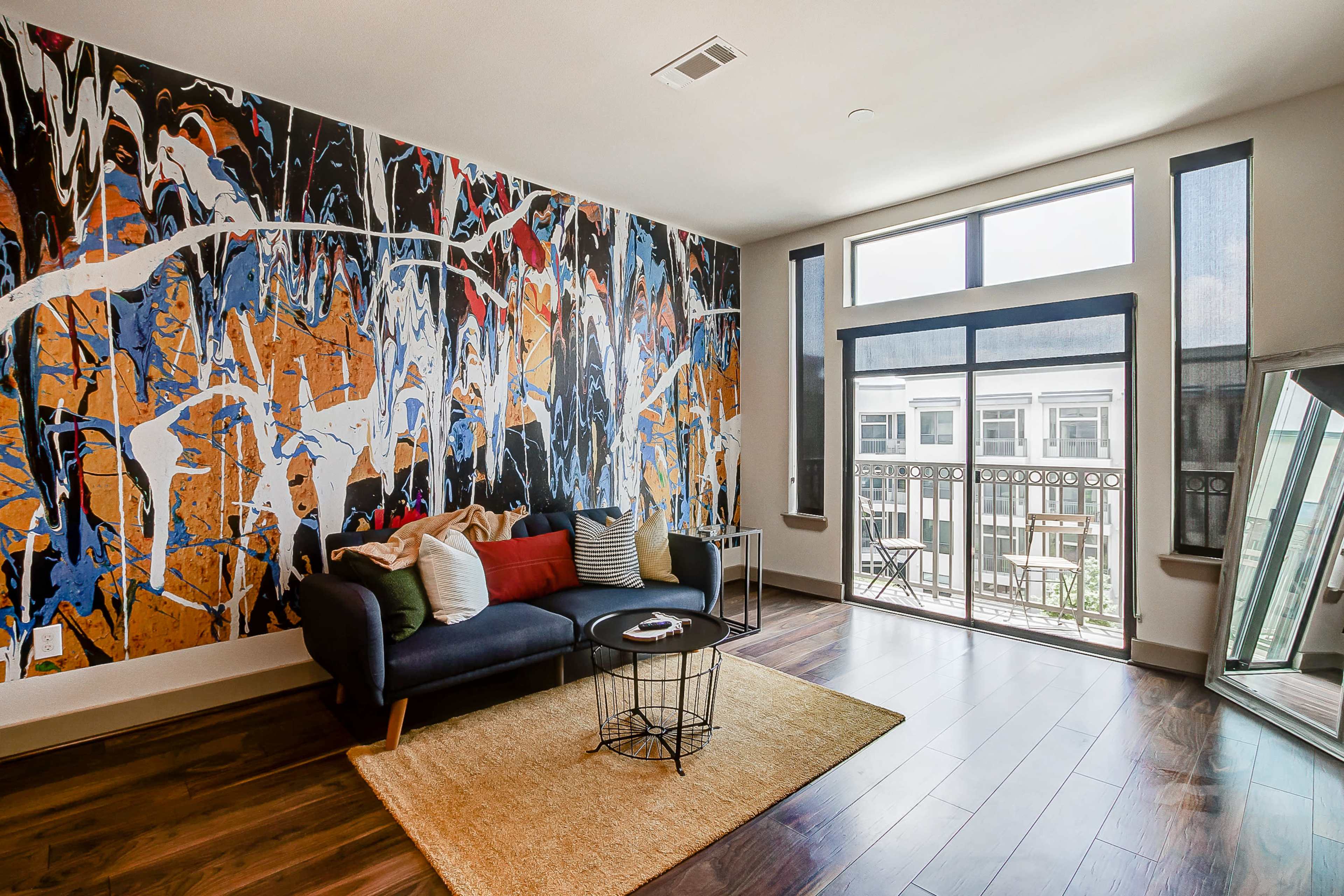 A living room with a large abstract mural on one wall, a dark sofa with colorful cushions, and a coffee table on a light rug, adjacent to a balcony visible through large windows.