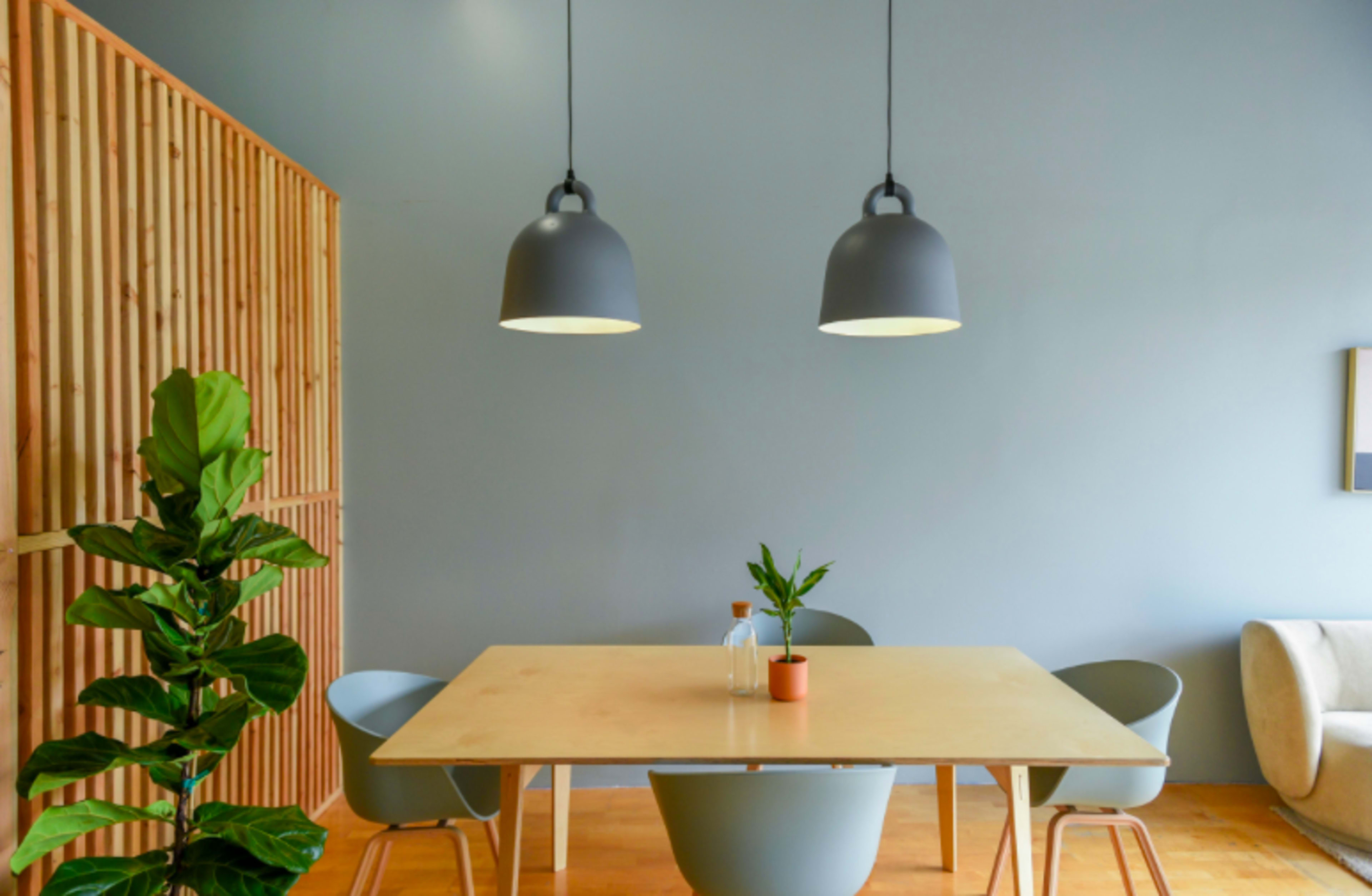 A modern dining area features a light wooden table surrounded by curved chairs, illuminated by two pendant lamps, with a potted plant in the corner.