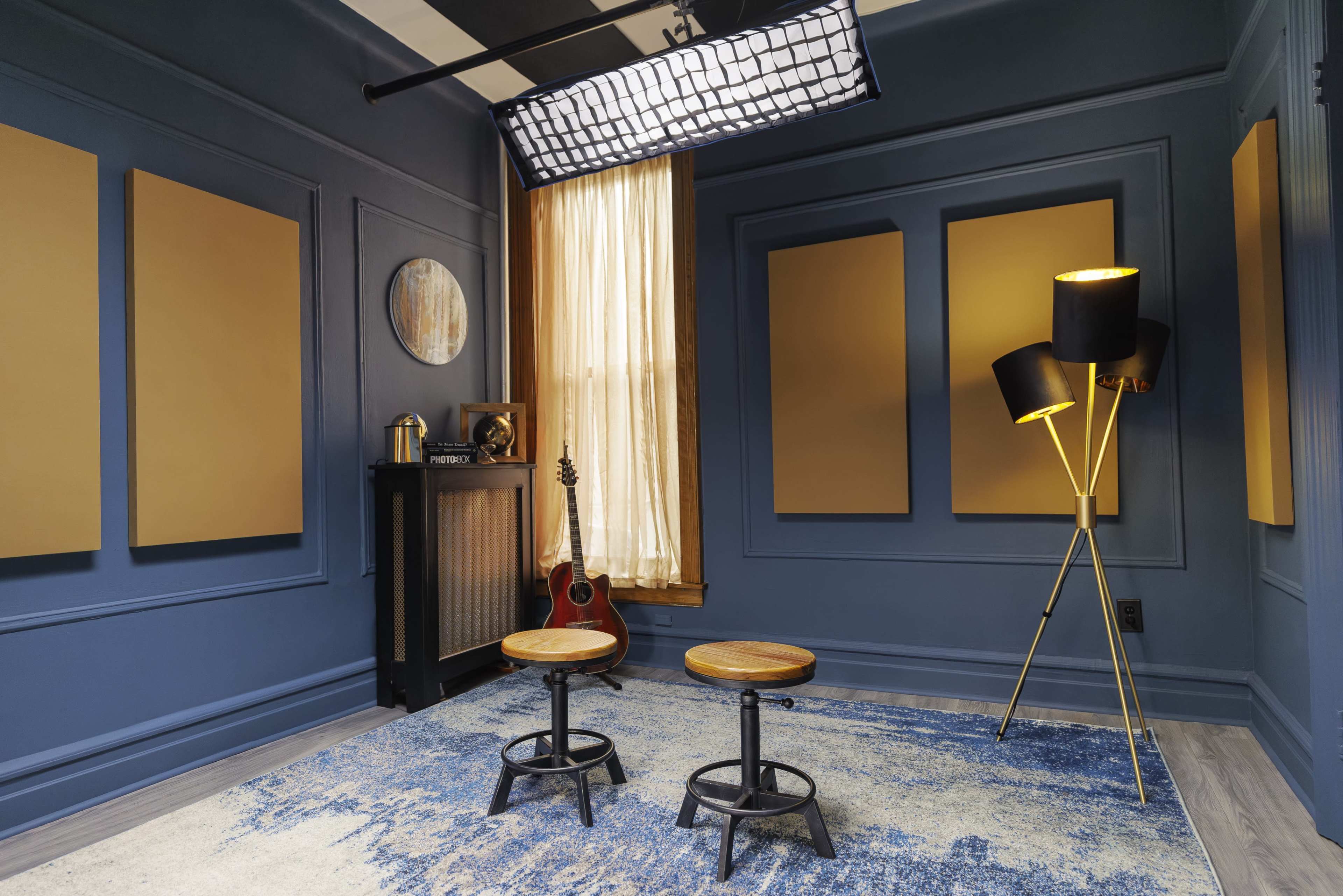 The image shows a styled room with blue walls, two wooden stools, a guitar, a tall lamp, and a large window with light filtering through.
