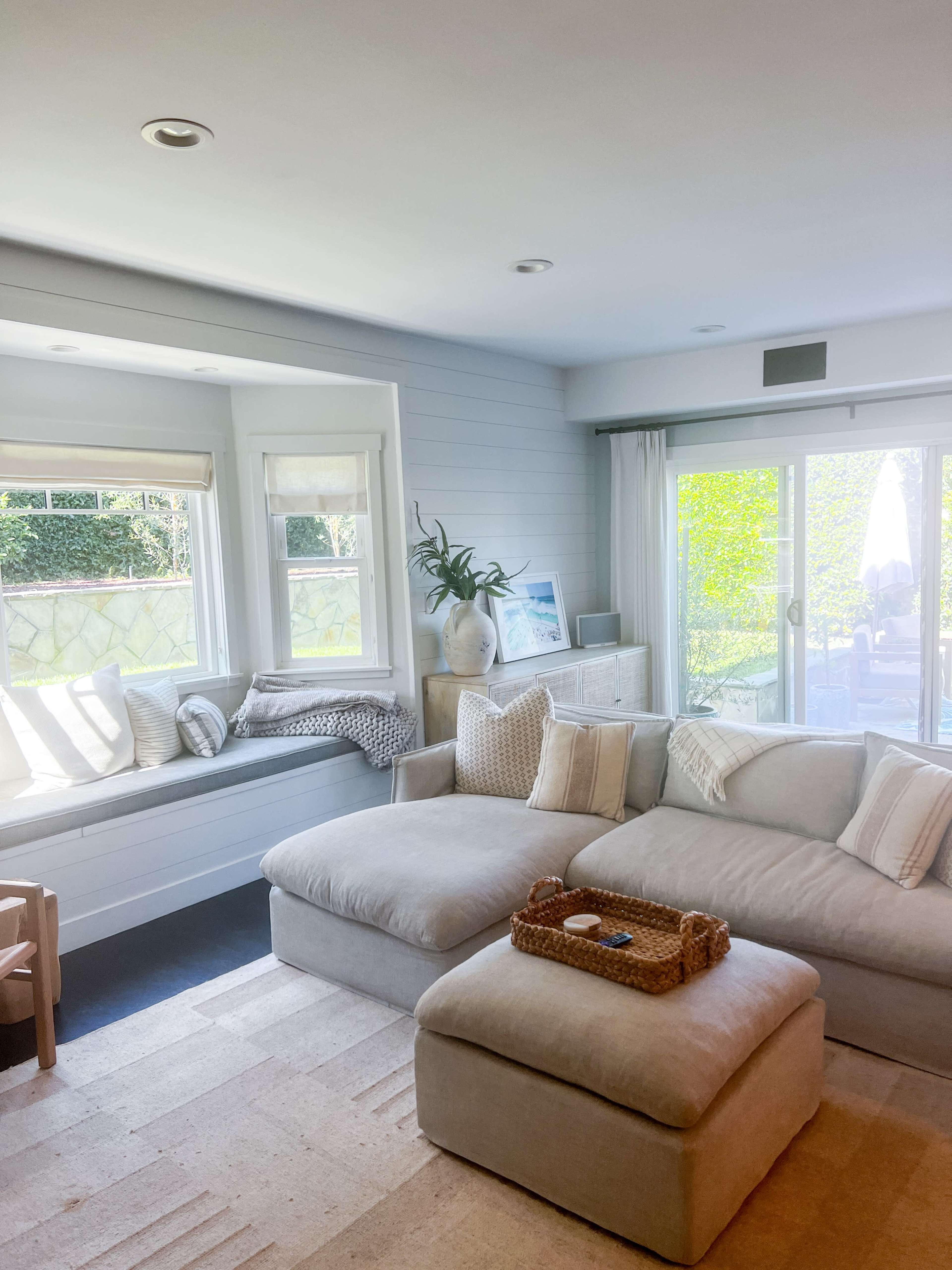 A light-filled living room features a sectional sofa, a window seat, and a view of an outdoor patio through glass doors.
