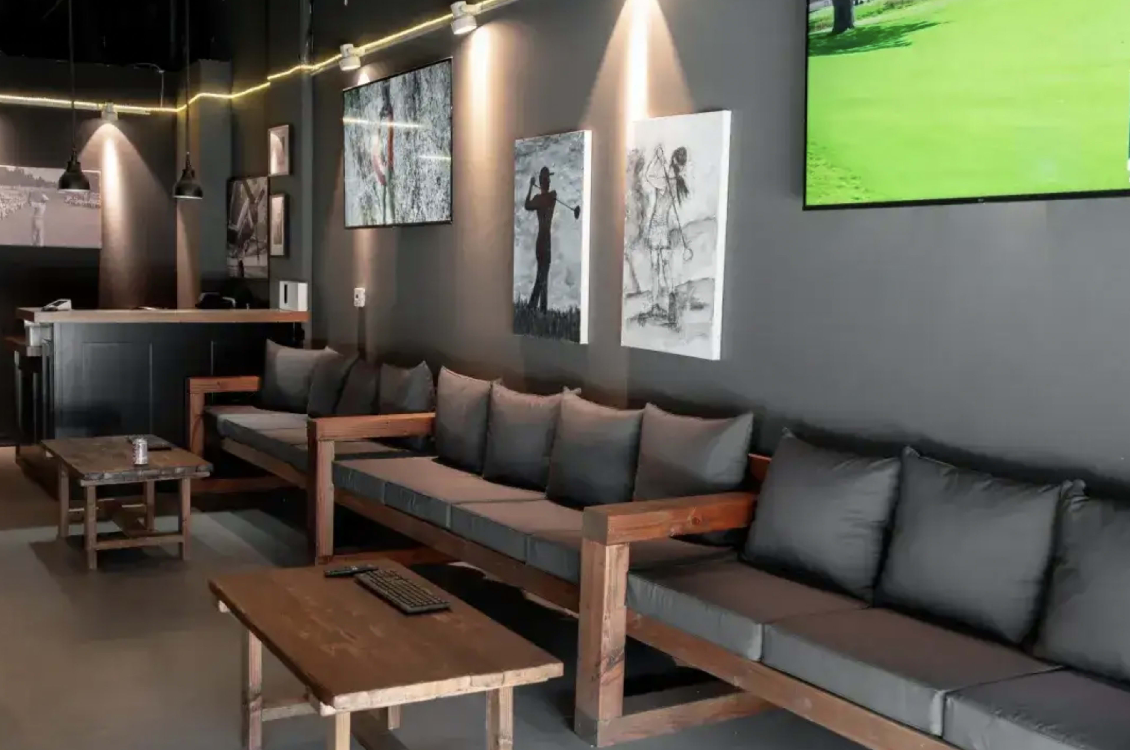 The image shows a modern lounge area featuring a long wooden sofa with gray cushions, a wooden coffee table, and wall-mounted televisions displaying a green landscape.