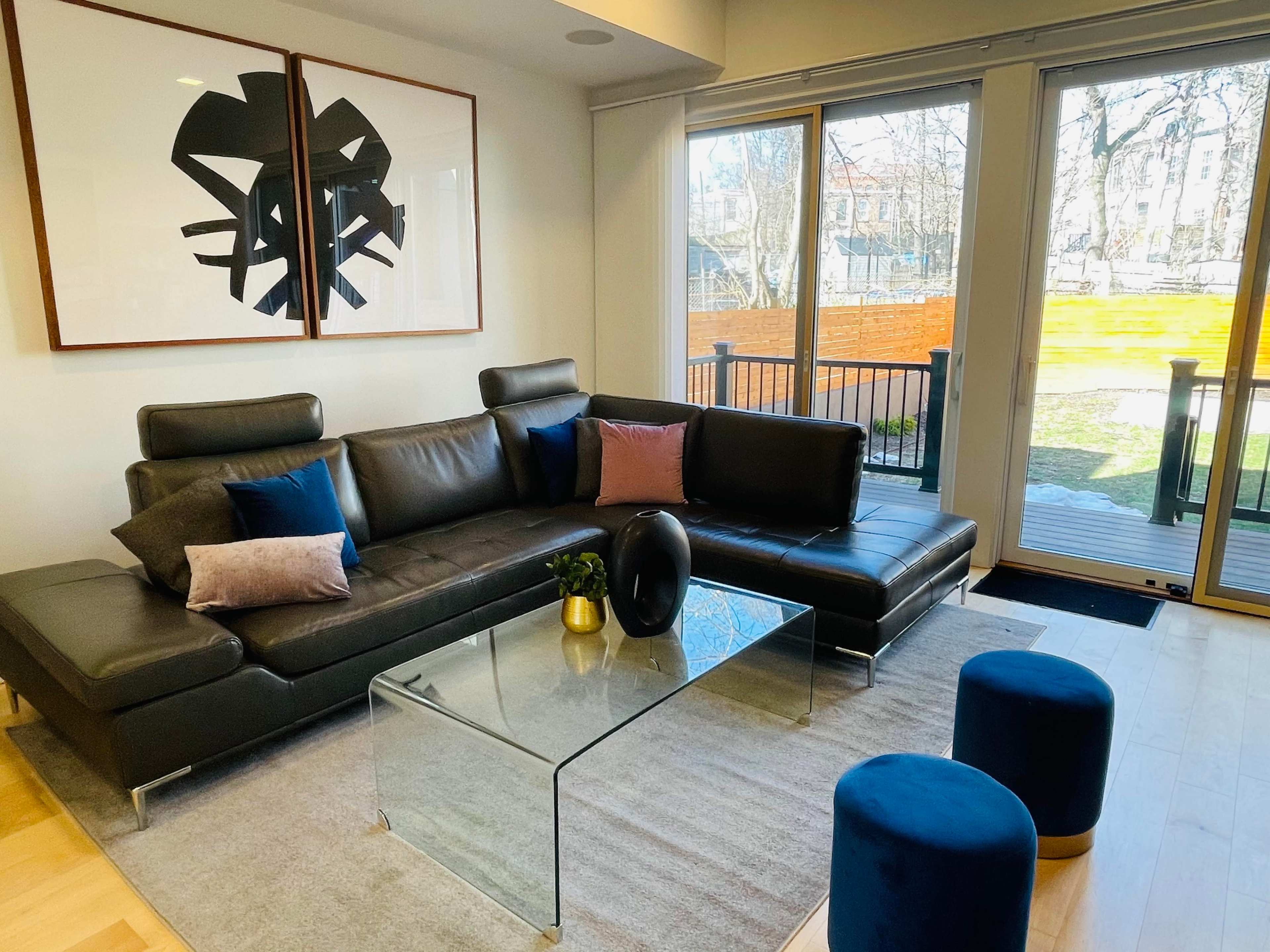 A modern living room featuring a black leather sectional sofa, a glass coffee table, and two blue ottomans, with large windows revealing an outdoor area.