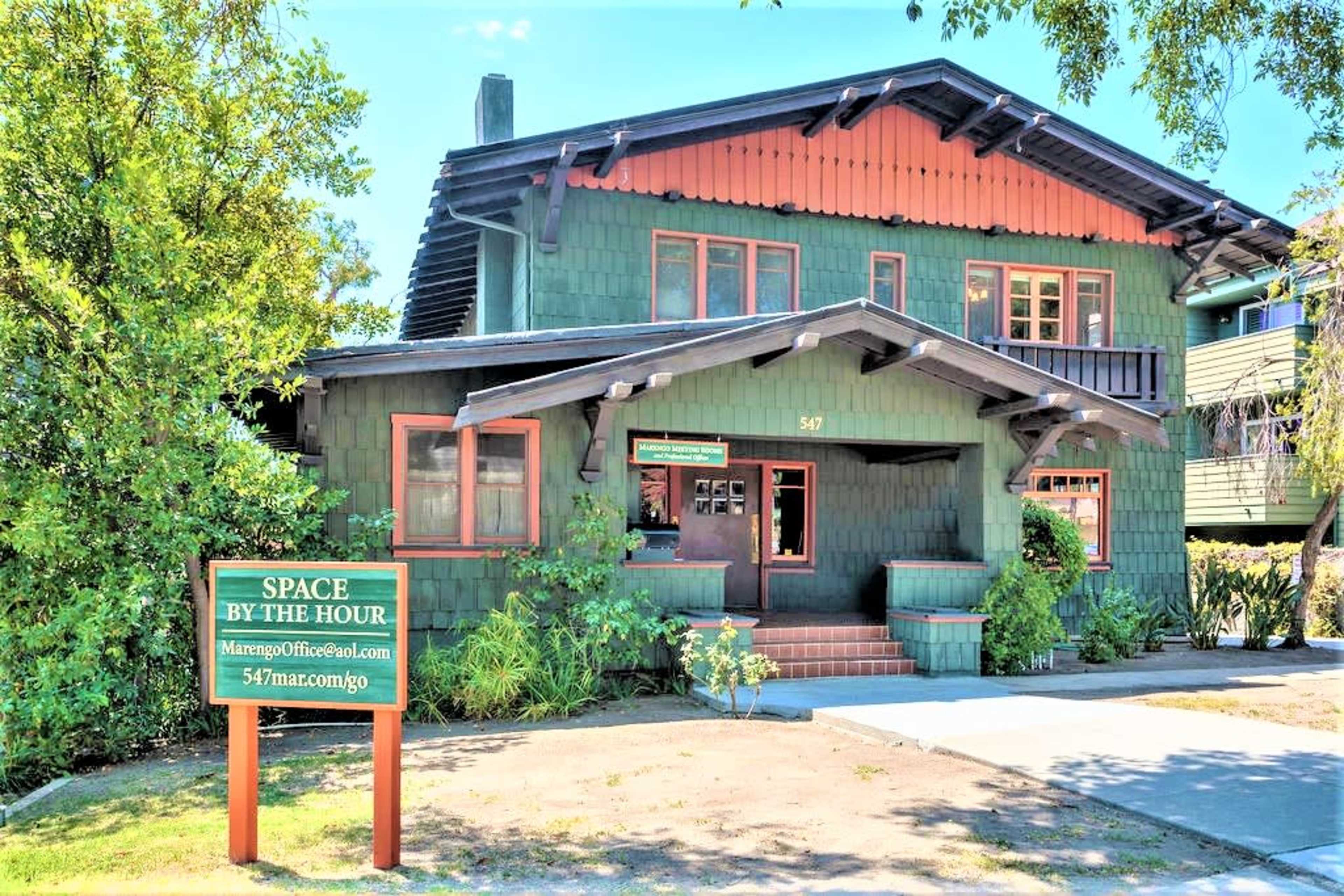 The image shows a green, two-story building with a distinctive architectural style, featuring large windows, a sign that reads "SPACE BY THE HOUR," and surrounded by greenery.