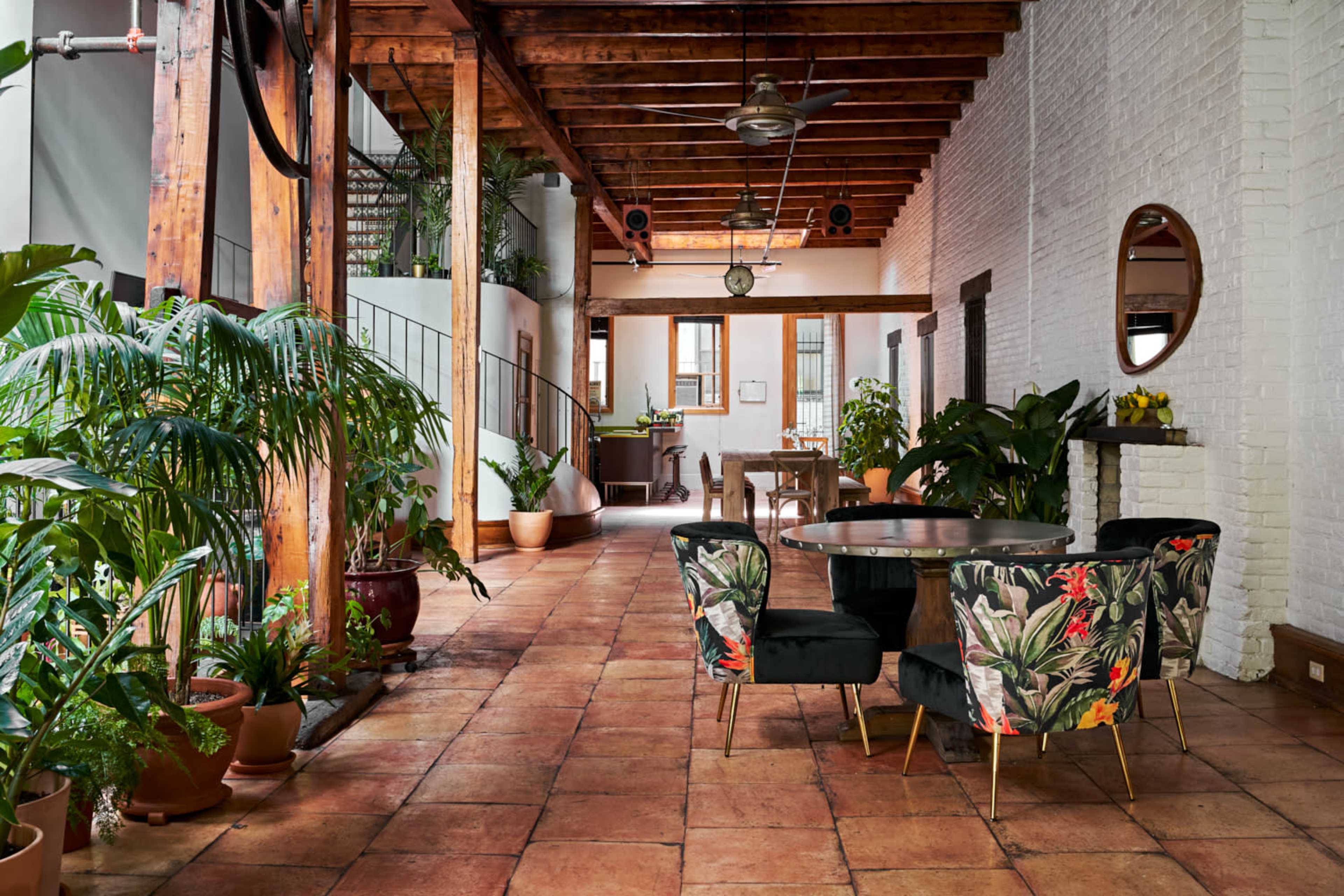 A spacious interior with terracotta tile flooring, a dining area featuring a round table and floral-patterned chairs, and large plants throughout the space.