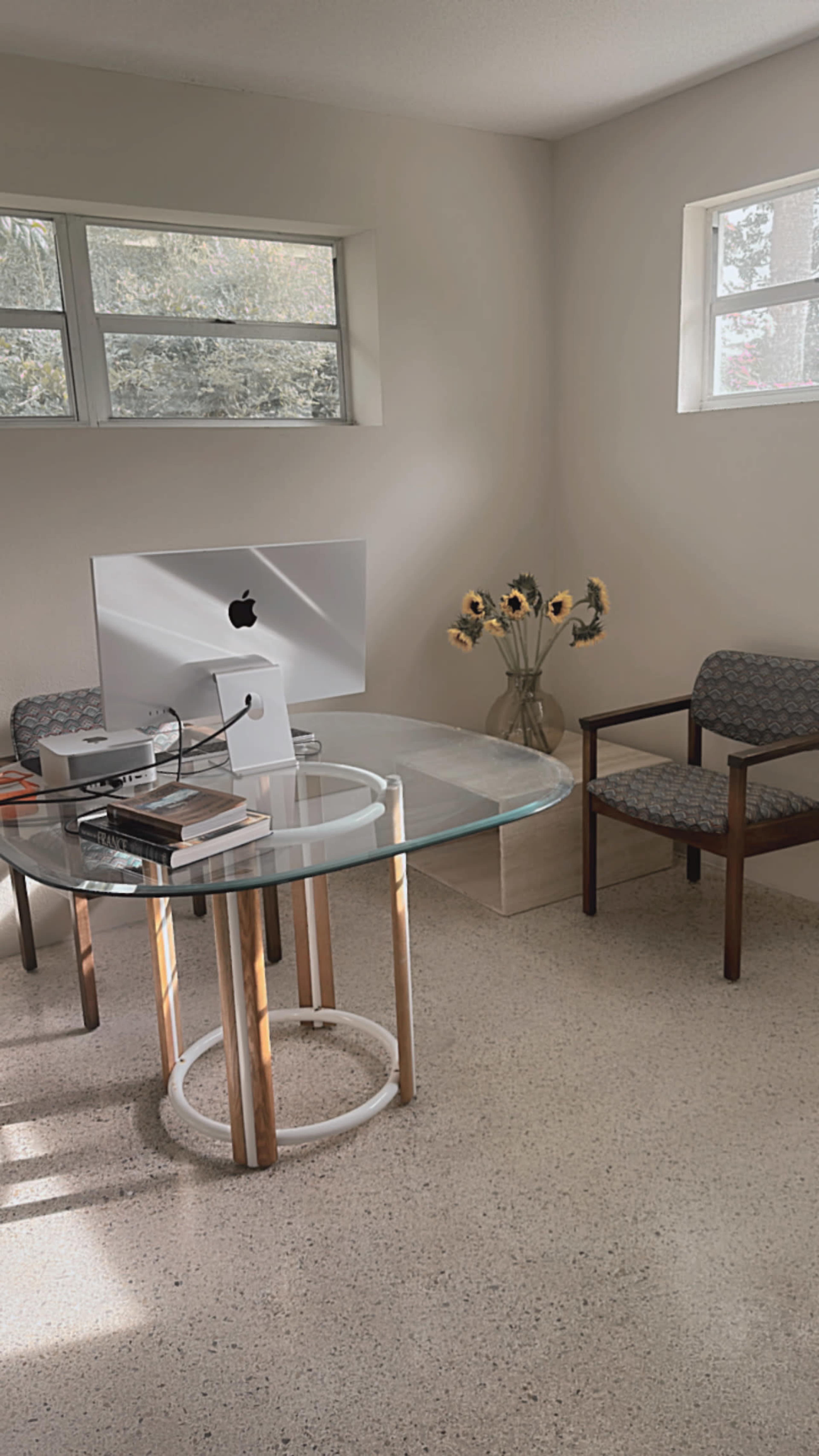 A minimalist office space with a glass table, an iMac computer, a stack of books, a chair, and a vase of sunflowers.