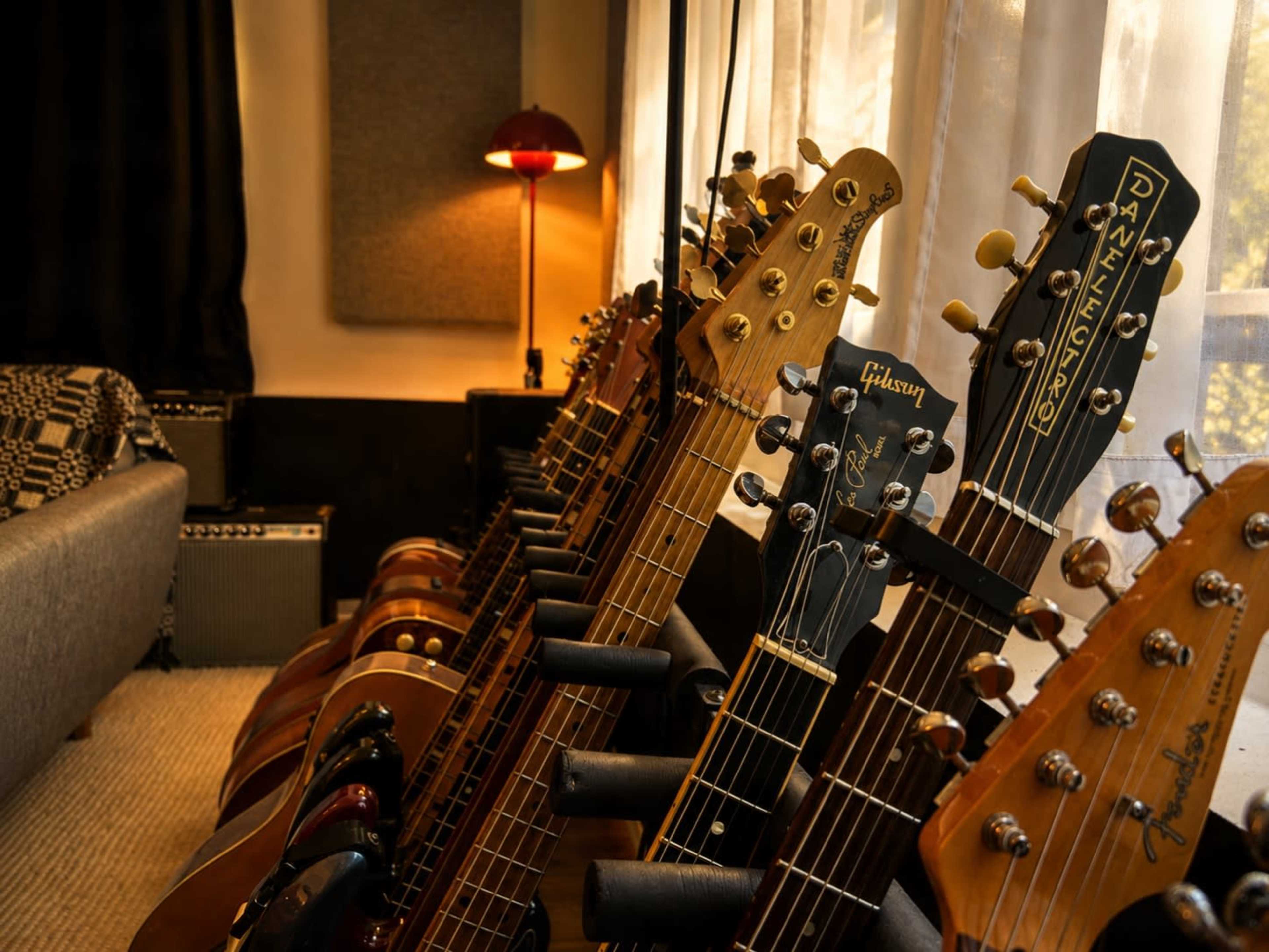 A row of various guitars is displayed on a rack in a well-lit room, with a lamp and a couch visible in the background.