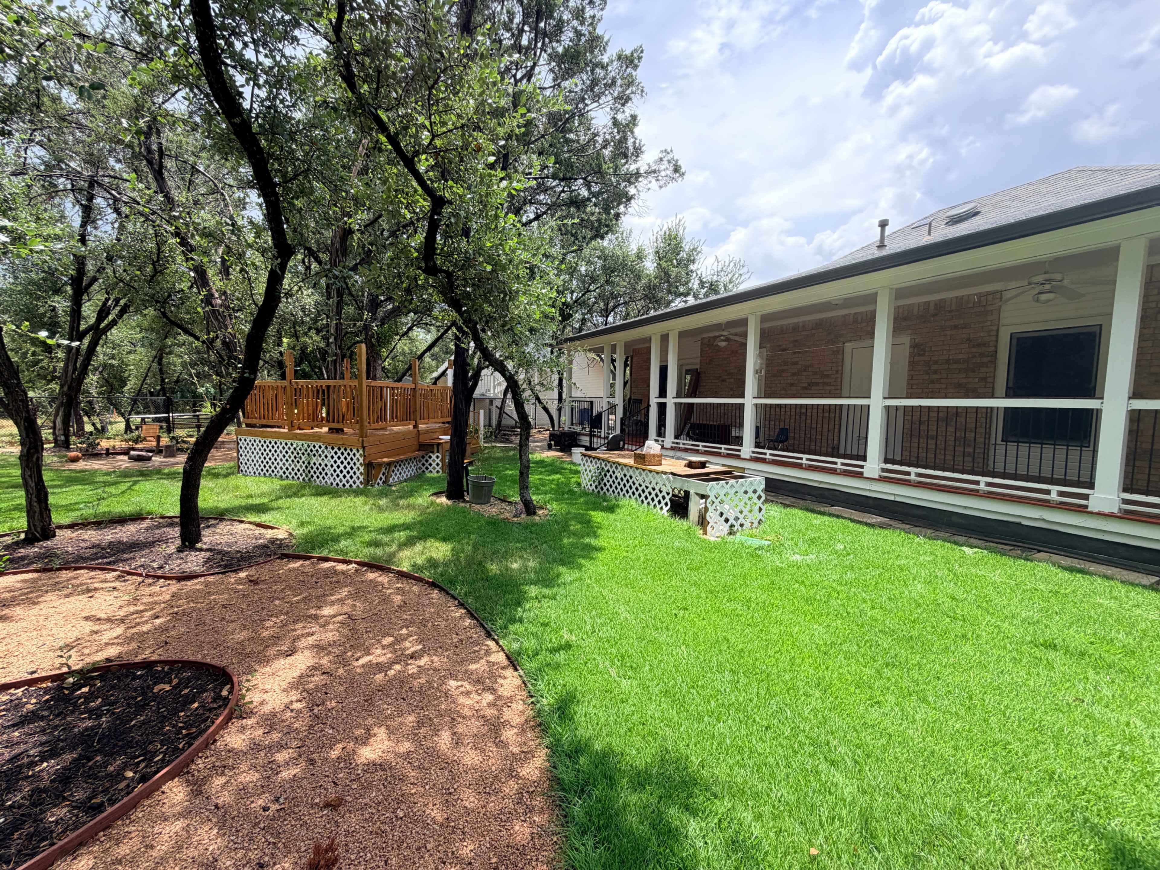 The image shows a landscaped backyard with a wooden deck, green grass, and trees surrounding a house.
