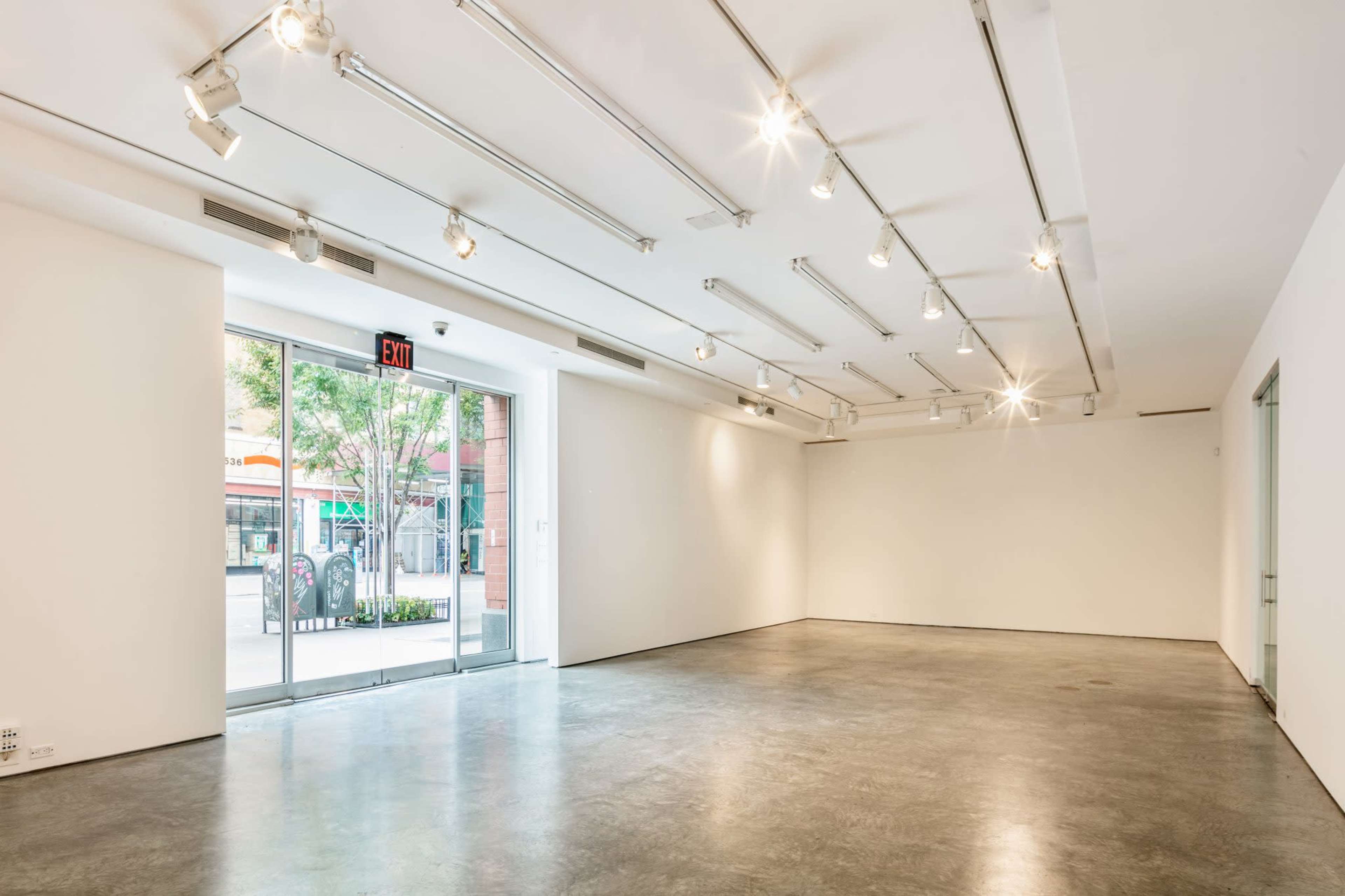 A spacious, well-lit gallery with white walls, concrete flooring, and large windows providing an exit to the outside.