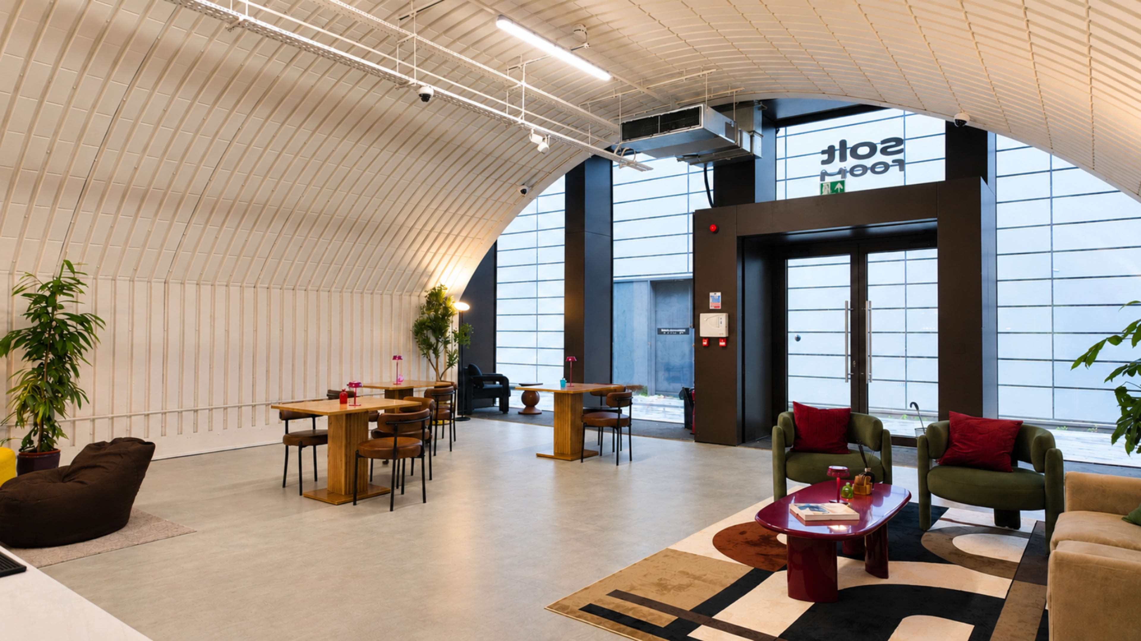 The image shows a modern indoor space with a curved ceiling, featuring seating areas and dining tables alongside large windows.