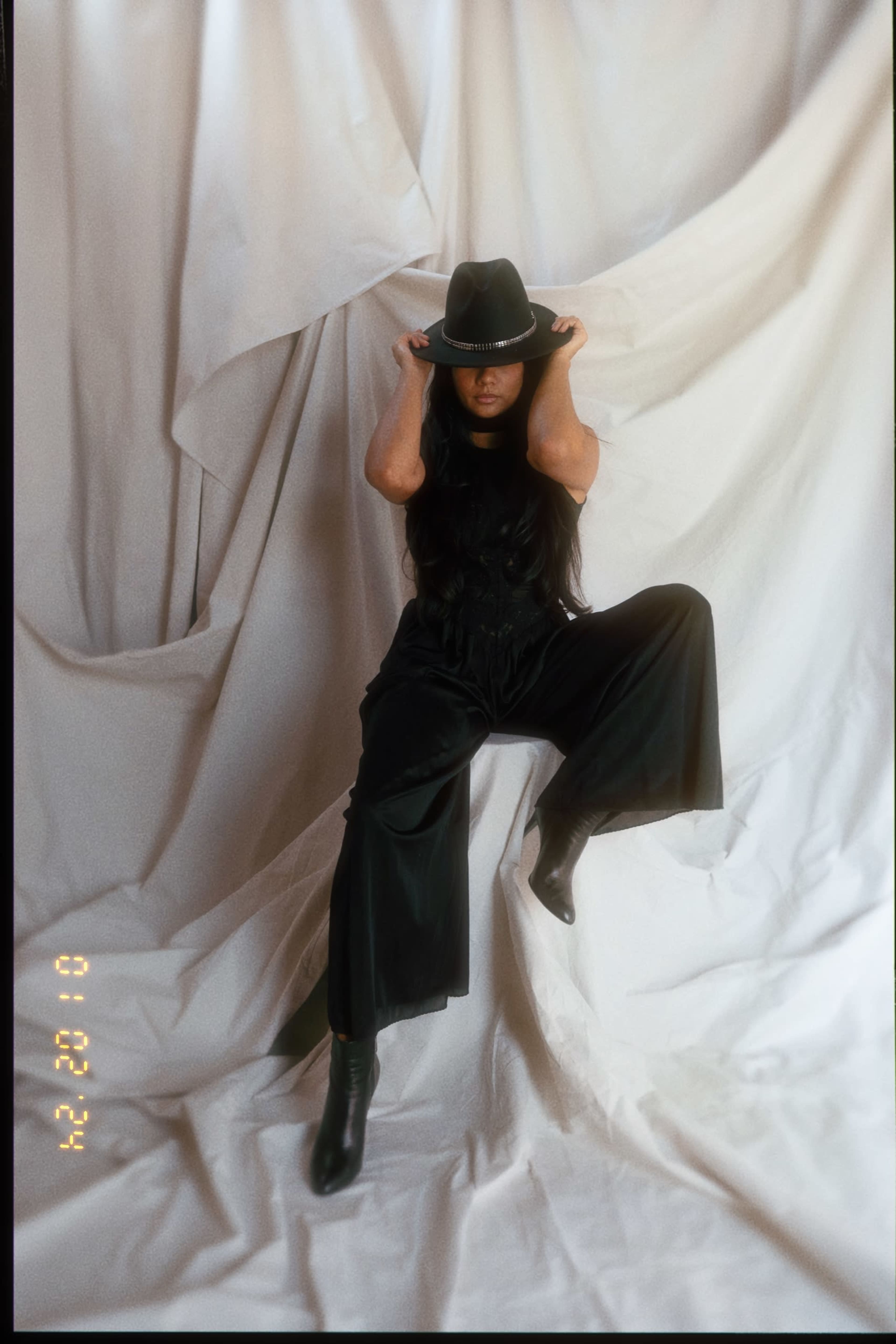 A person wearing a black hat and outfit poses on a draped white background, one hand on their head and legs apart.