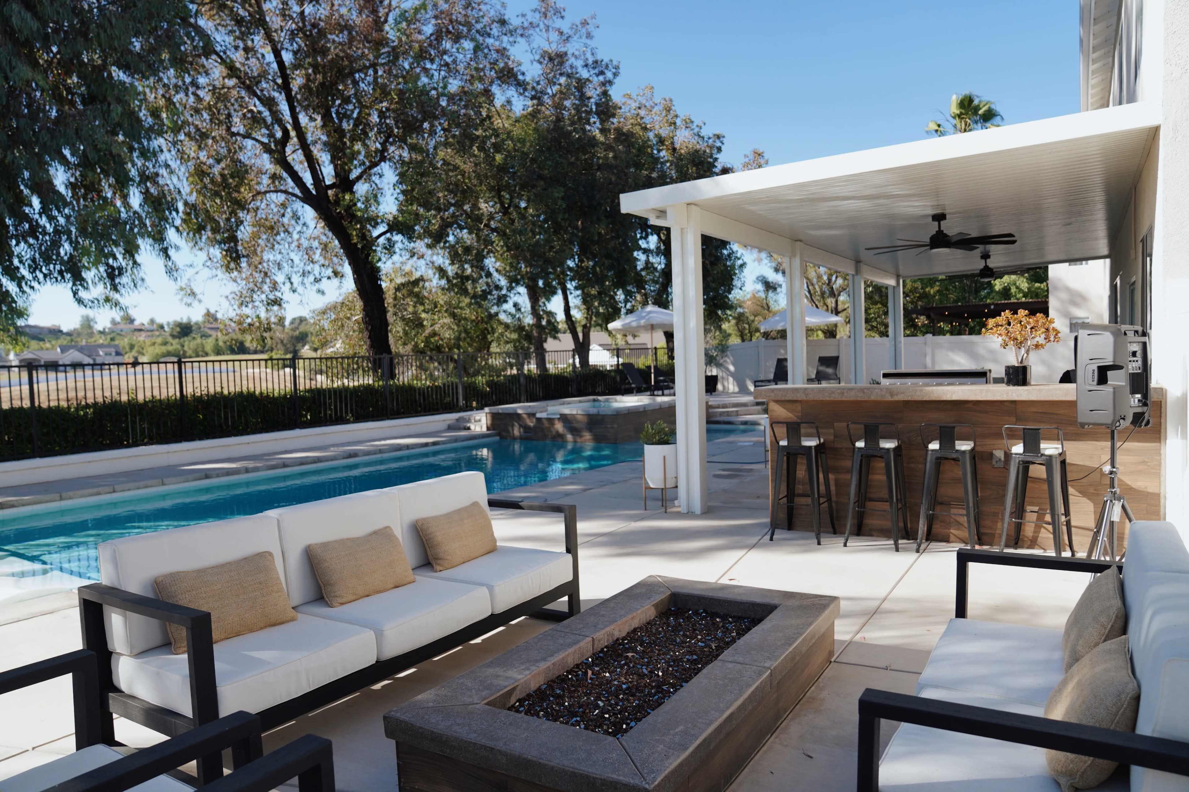 A modern outdoor living space features a seating area with white couches and a fire pit, overlooking a pool and a covered bar area.