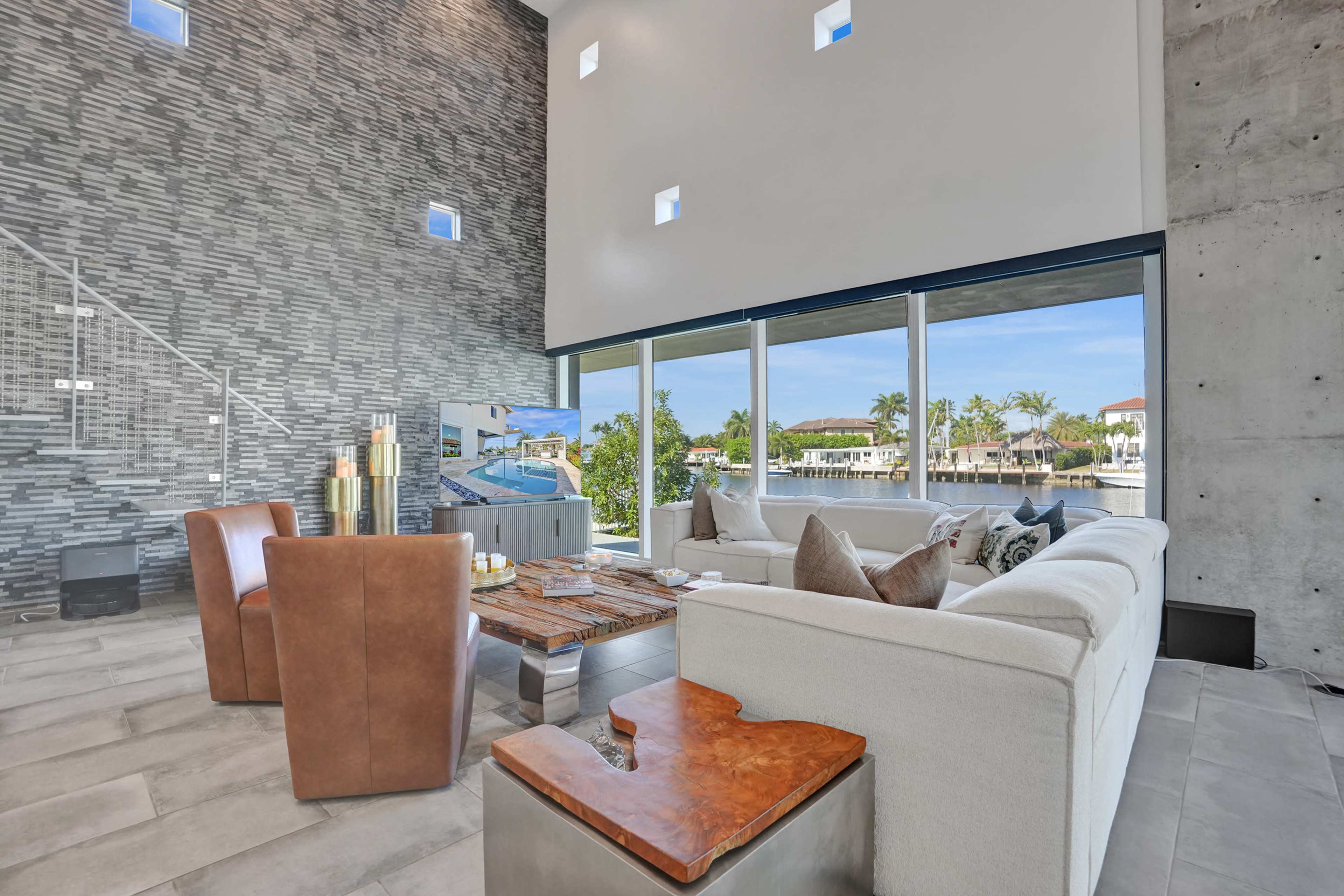 The image depicts a modern living area with large windows overlooking a waterfront scene, featuring minimalist furniture and a textured stone wall.