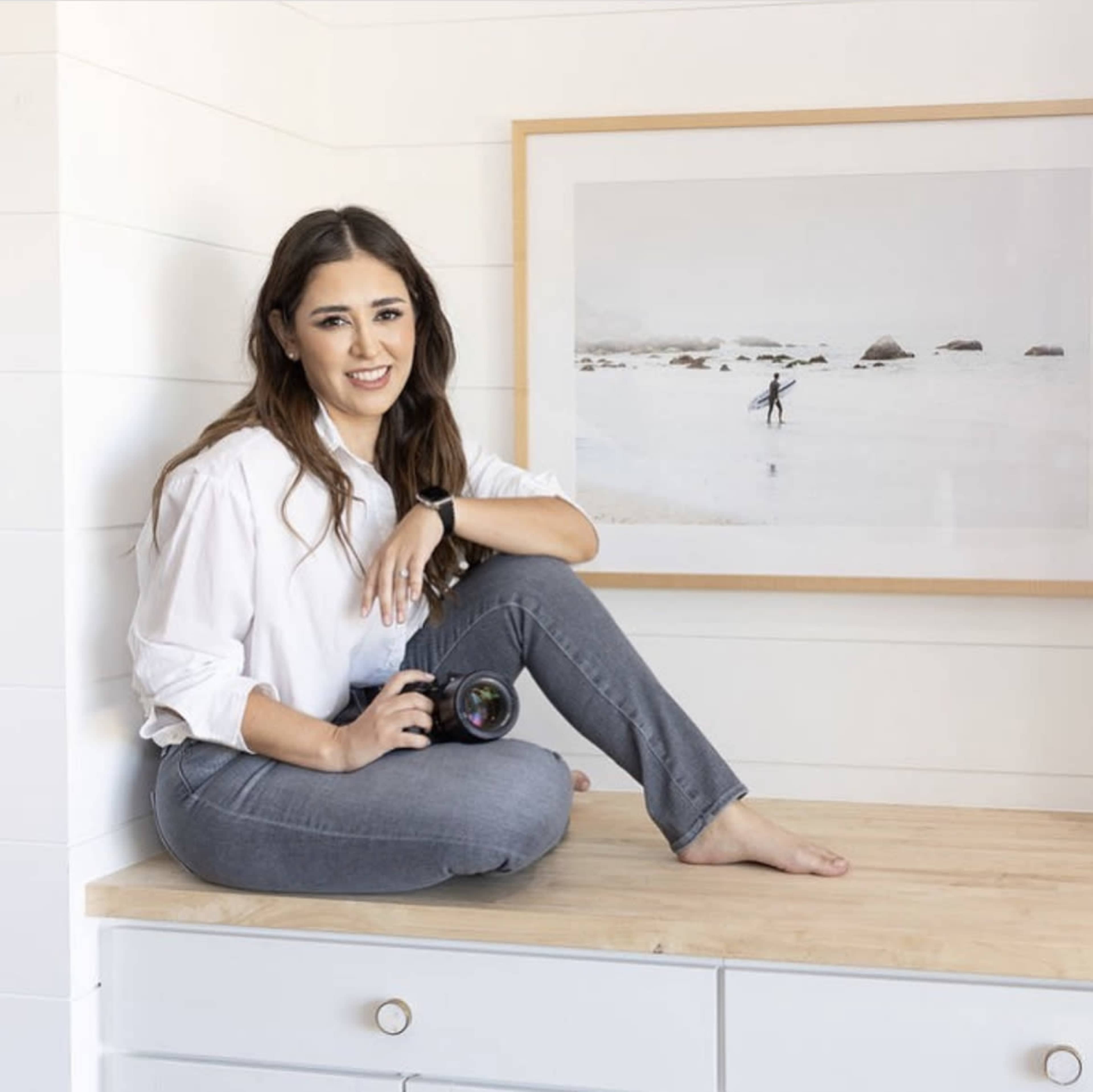 A woman is sitting cross-legged on a wooden surface, holding a camera, next to a framed photograph of a person surfing on a beach.