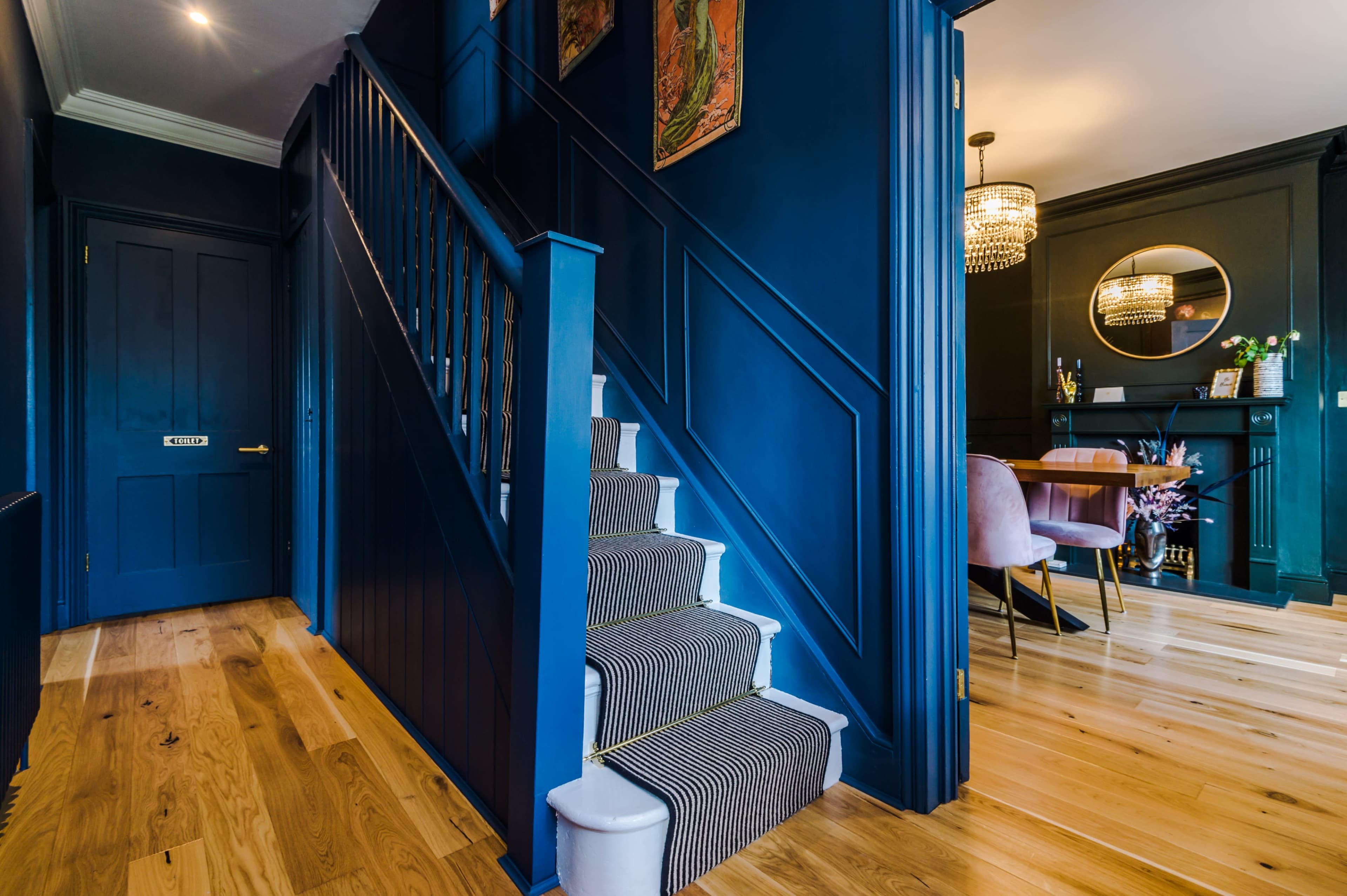 A stairway with a dark blue wall and wooden flooring, leading to a dining area in the background with a round mirror and modern light fixture.