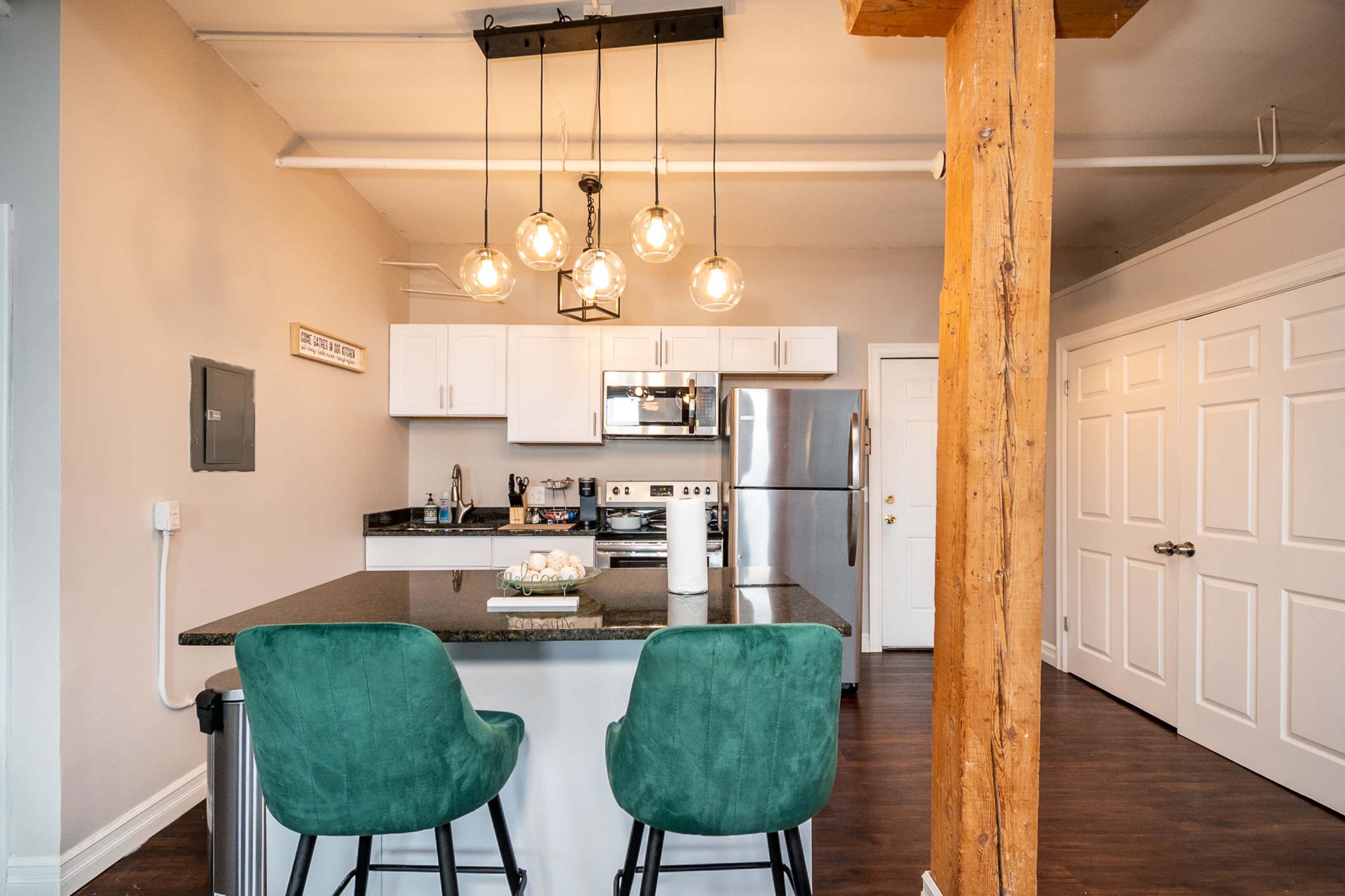 A modern kitchen with a large island featuring two green bar stools, stainless steel appliances, and overhead pendant lighting.