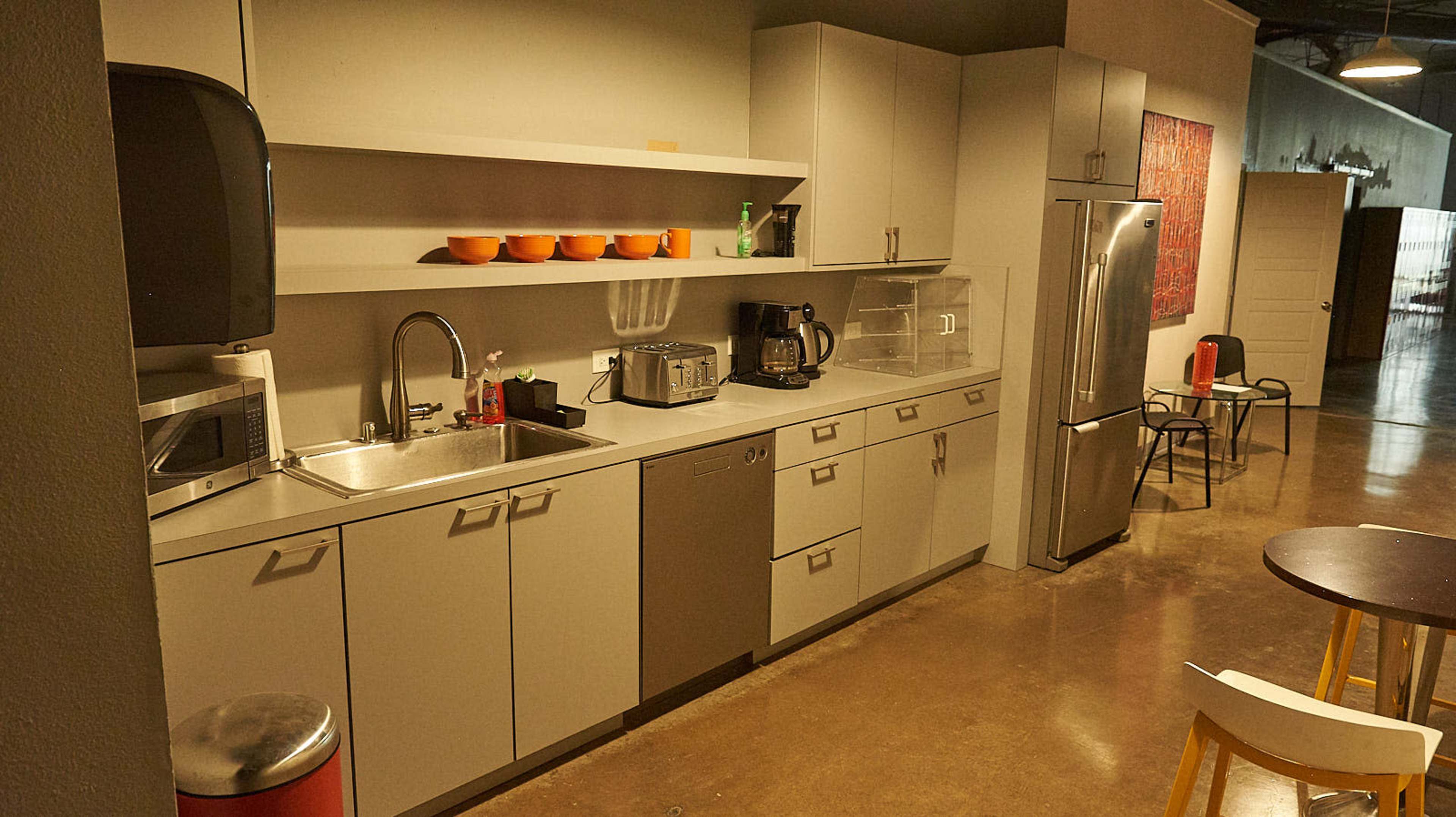 A modern kitchen with stainless steel appliances, including a refrigerator, microwave, and coffee maker, against a backdrop of light gray cabinetry and a concrete floor.