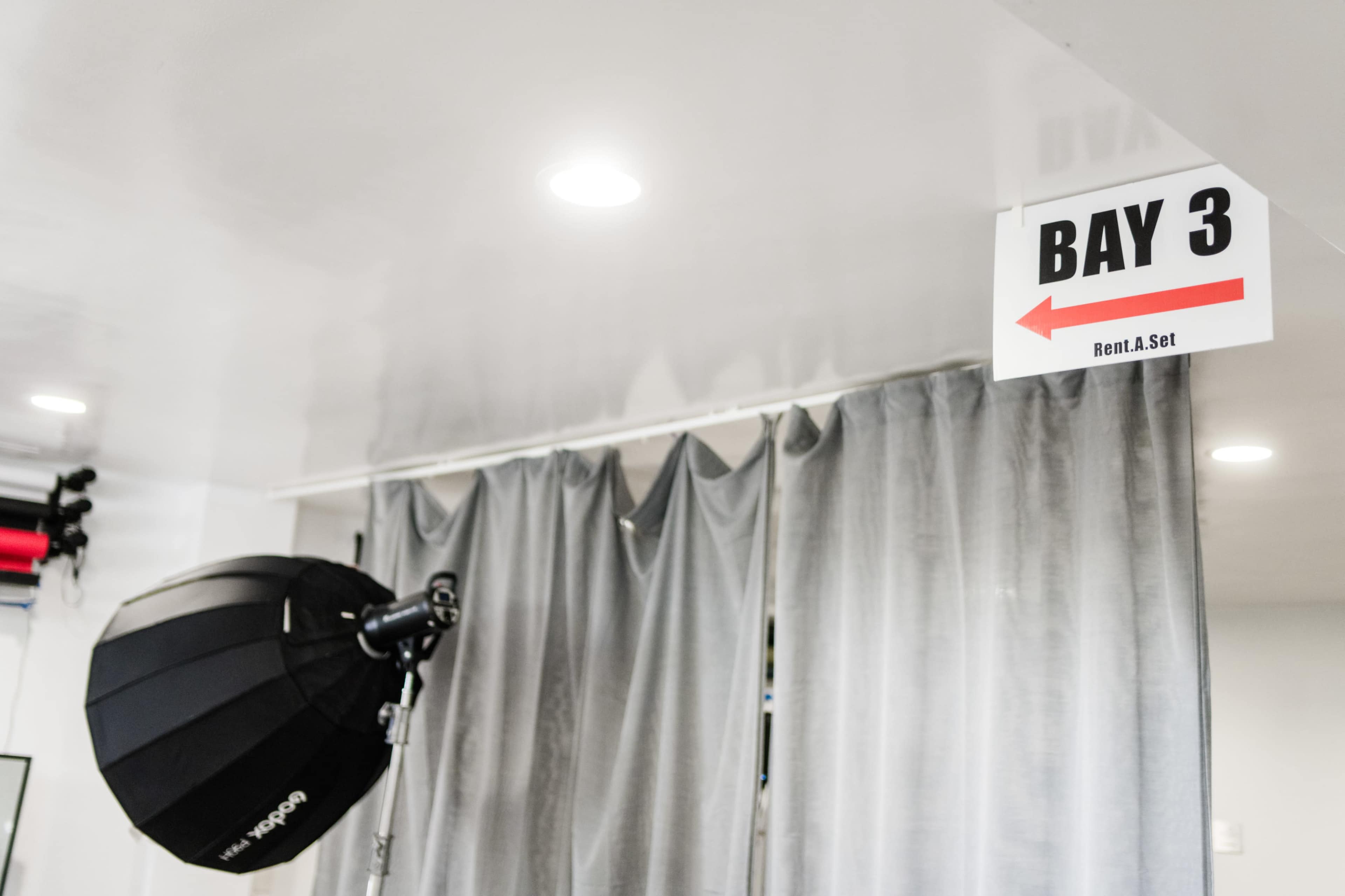 A sign labeled "BAY 3" with a red arrow points towards a section draped with gray curtains in a studio space.