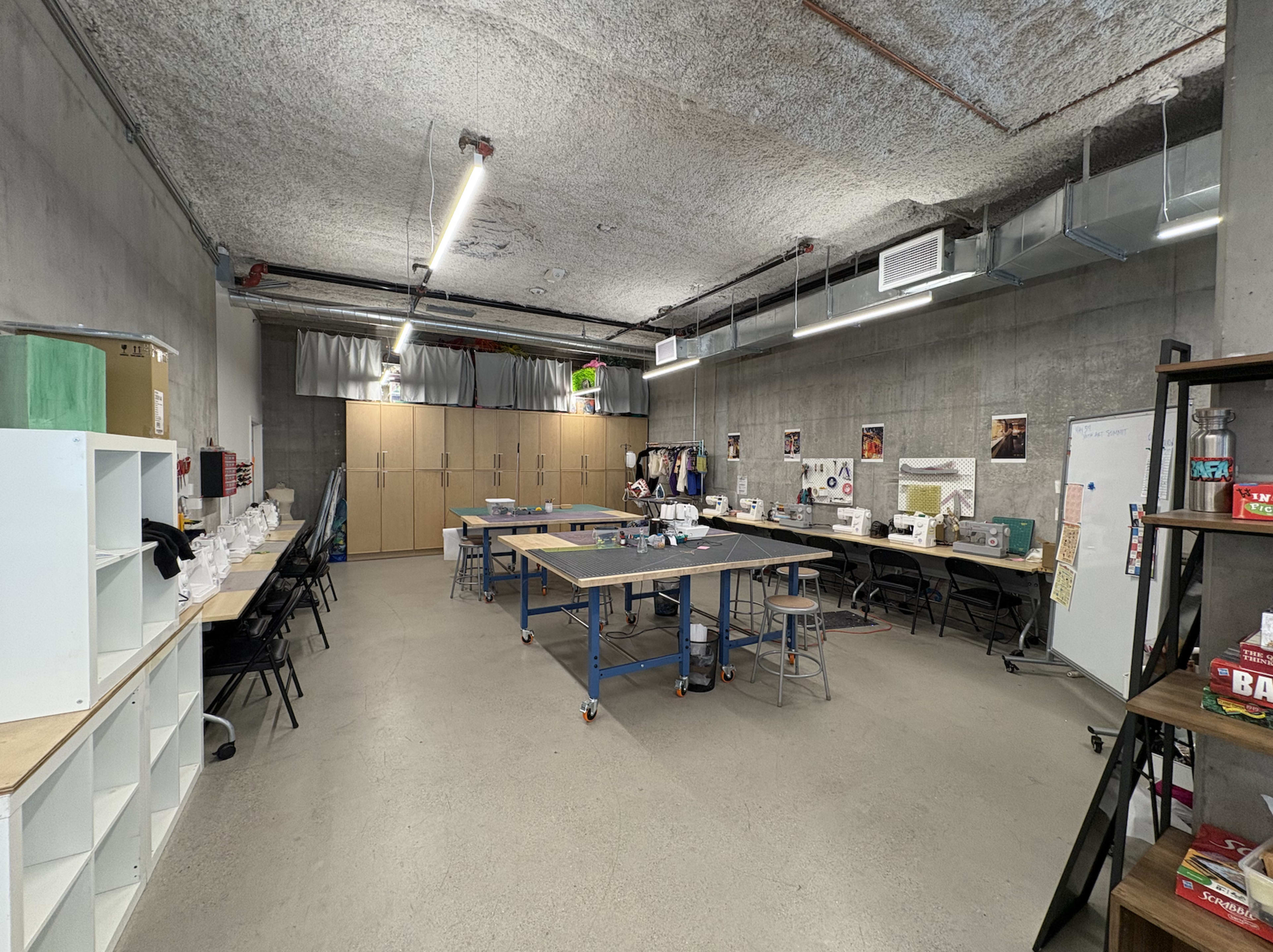 The image shows a spacious workshop with various tables and chairs, organized shelving units, and unfinished concrete walls.