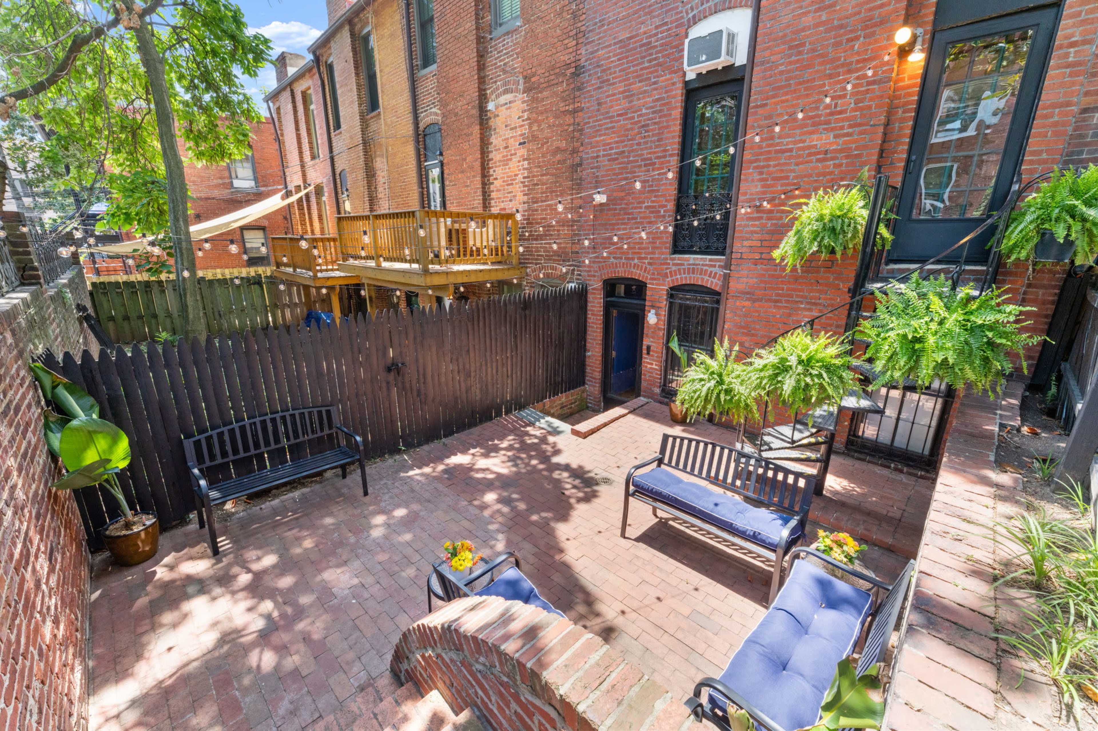 The image shows a brick-paved courtyard with seating, surrounded by brick buildings and greenery, including potted plants and wooden decking.