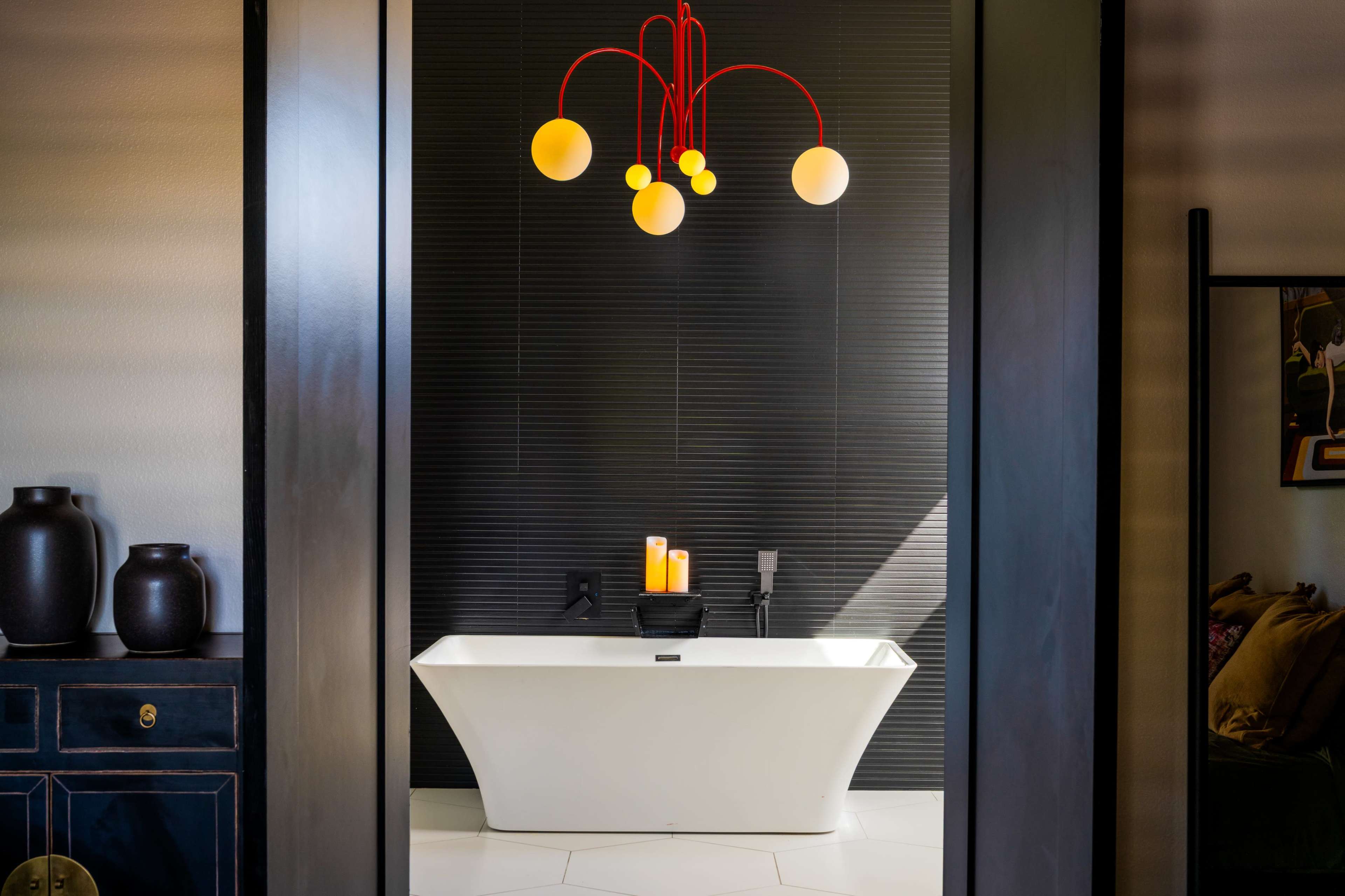 A modern bathroom features a freestanding white bathtub under a colorful pendant light against a dark tiled wall.