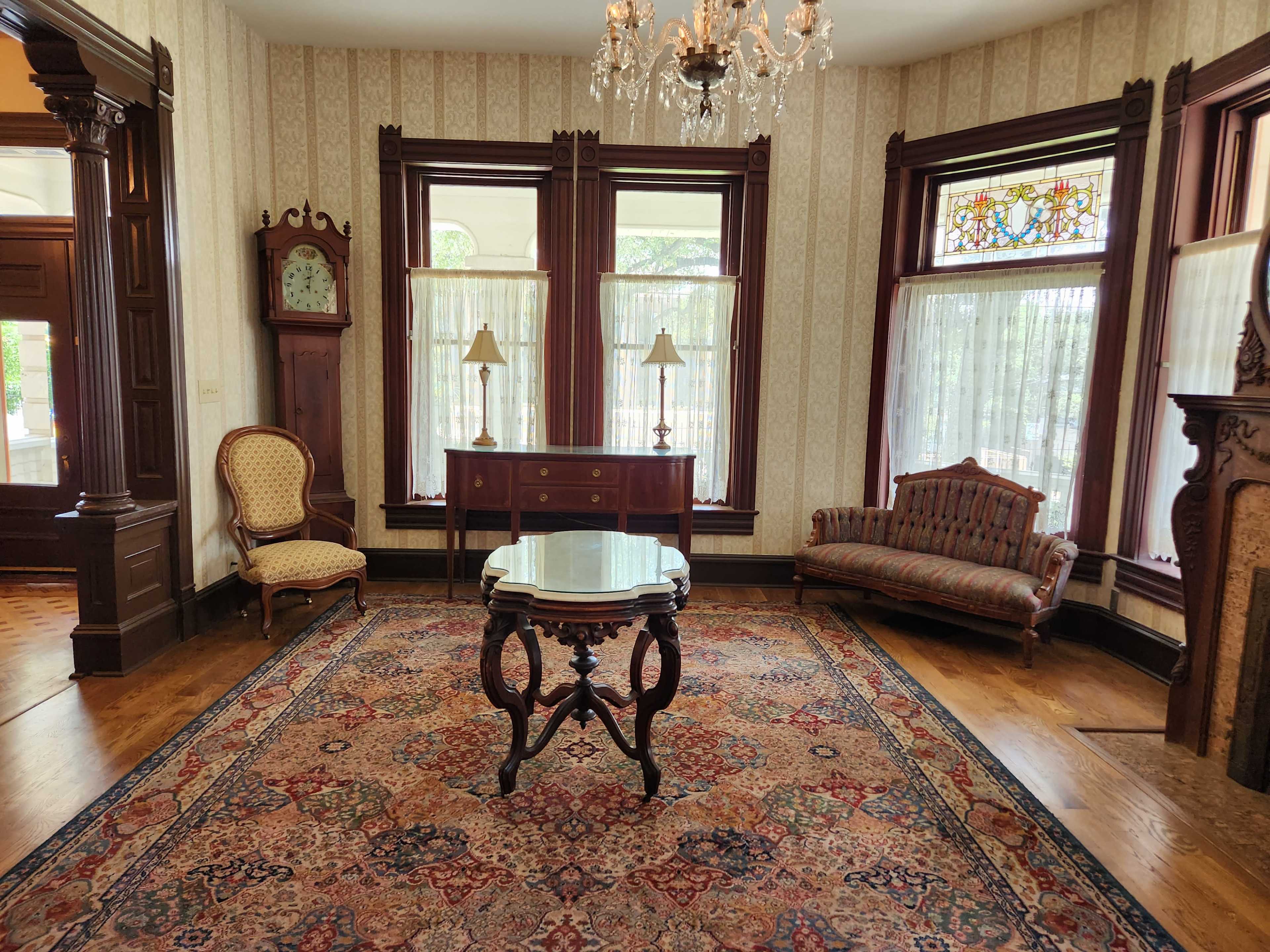 The image features a vintage sitting room with ornate woodwork, a central table, and a patterned rug, illuminated by natural light through large windows.
