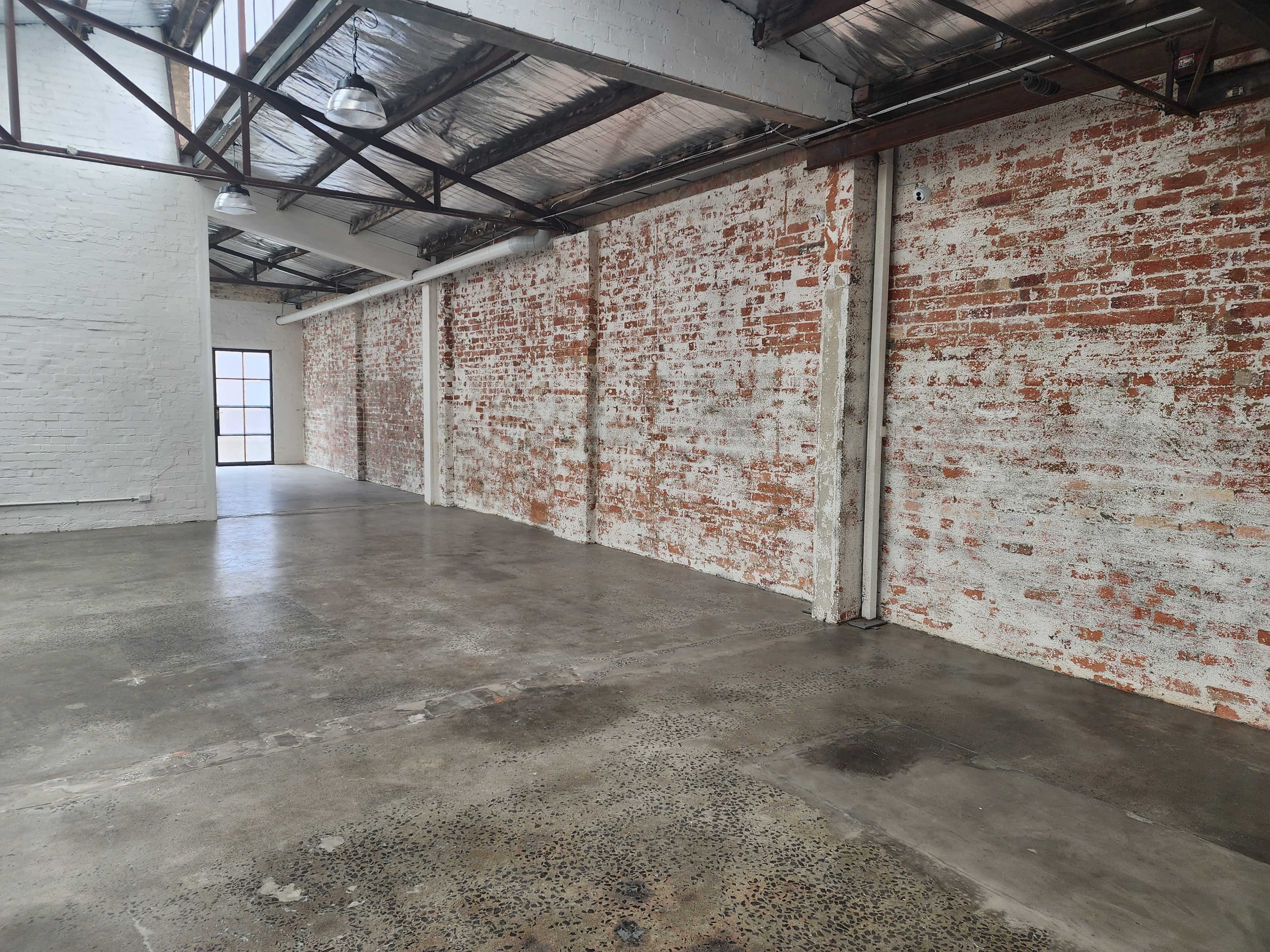 The image shows a spacious industrial room with bare brick walls and a concrete floor, illuminated by natural light from large windows.