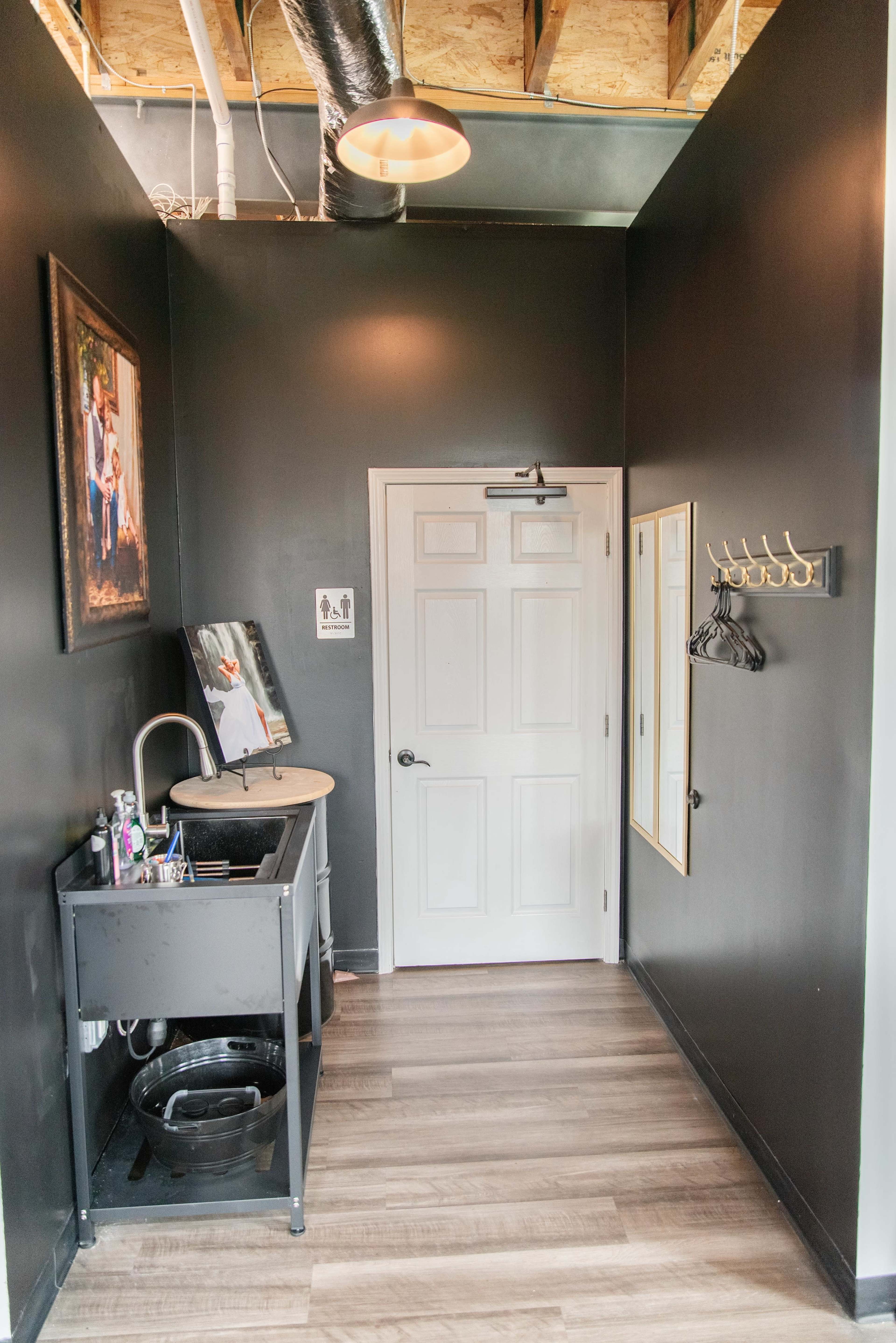 The image shows a narrow hallway with black walls, featuring a sink and countertop on the left, a door ahead, a mirror on the right, and a coat rack on the wall.