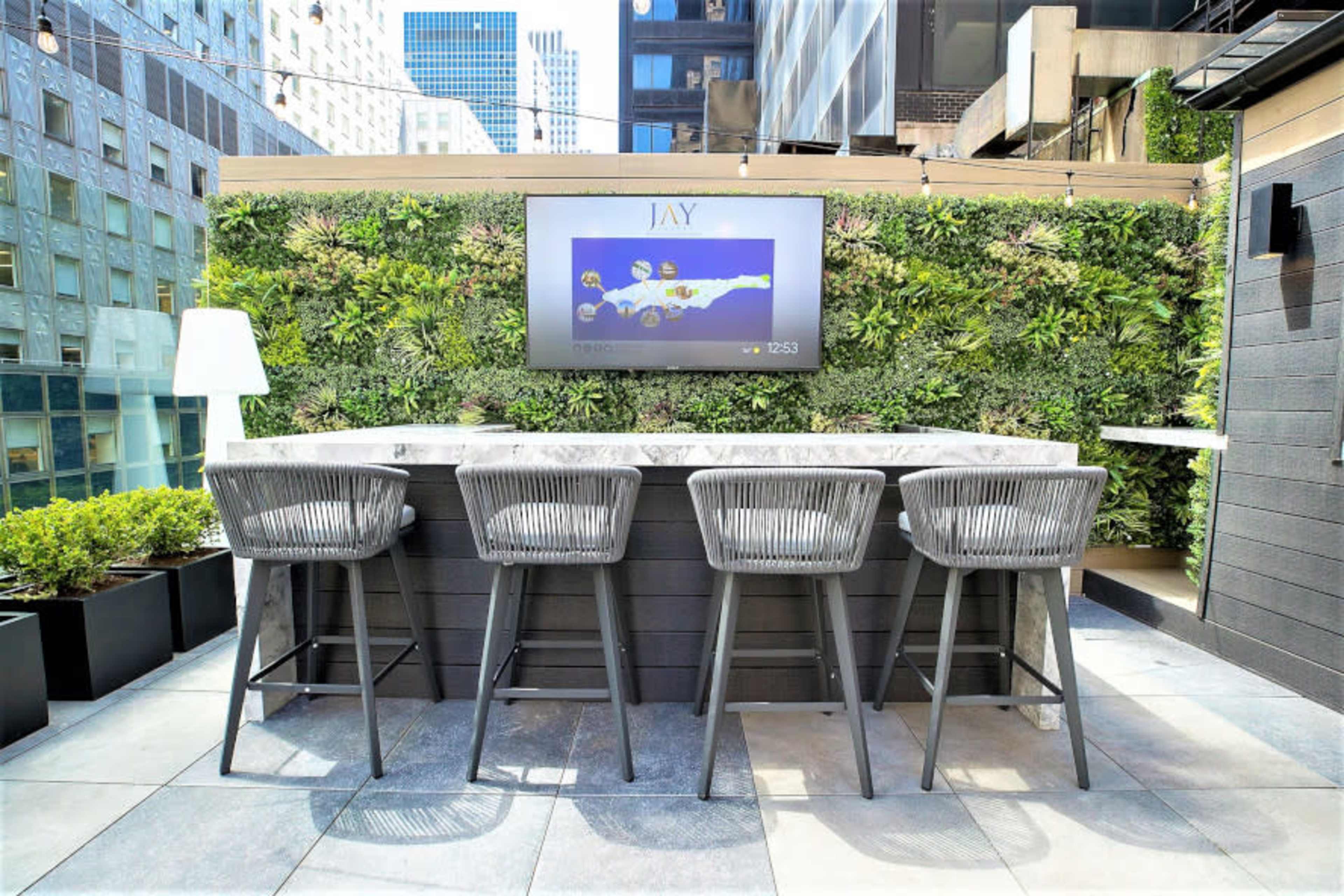 The image shows a modern rooftop bar area with a stone counter and four high chairs, surrounded by green wall planters and featuring a large flat-screen TV.