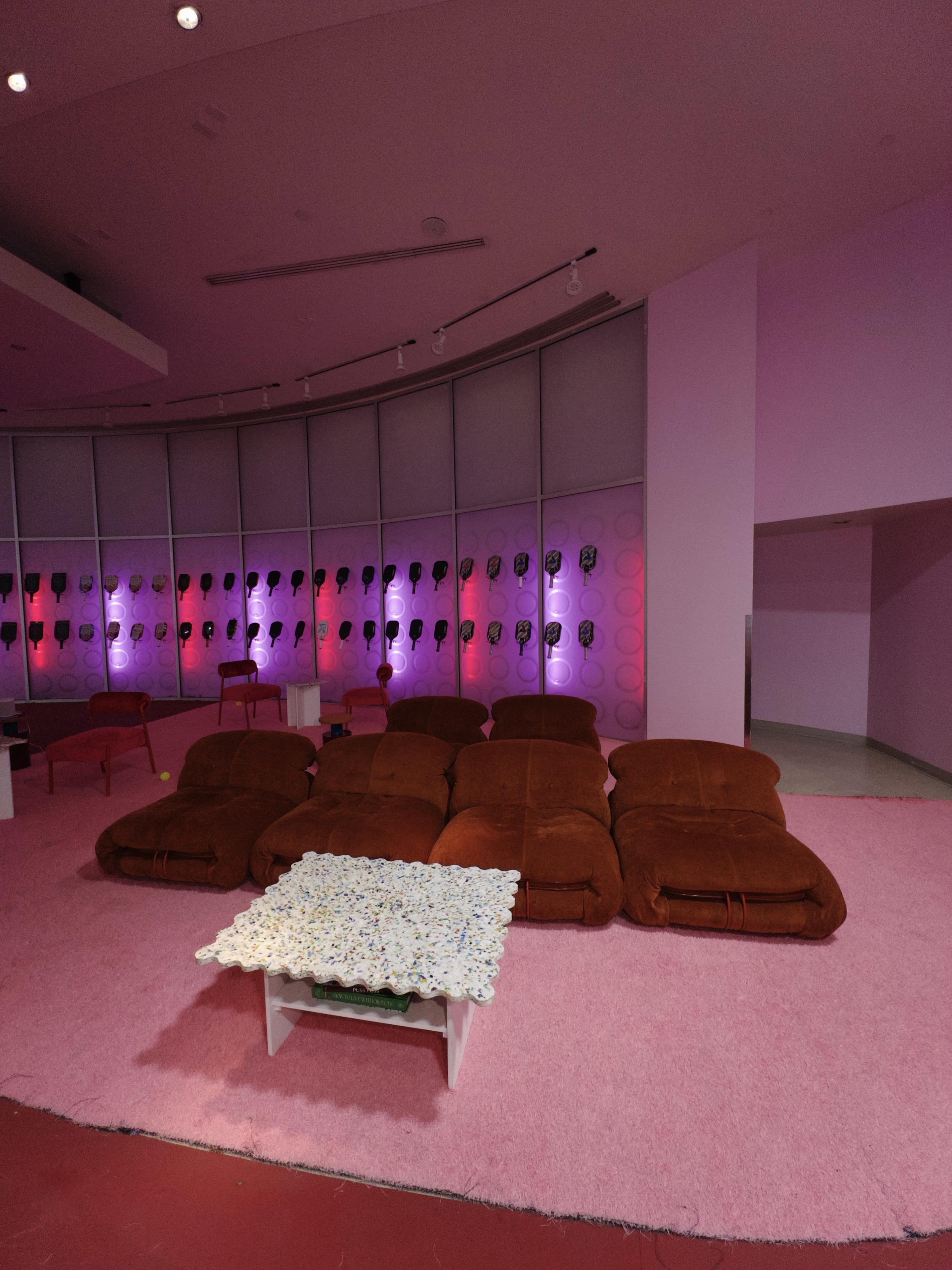 A seating area with brown sofas arranged in a U-shape, a white coffee table with a floral centerpiece, and a backdrop of lockers illuminated by pink and purple lights.