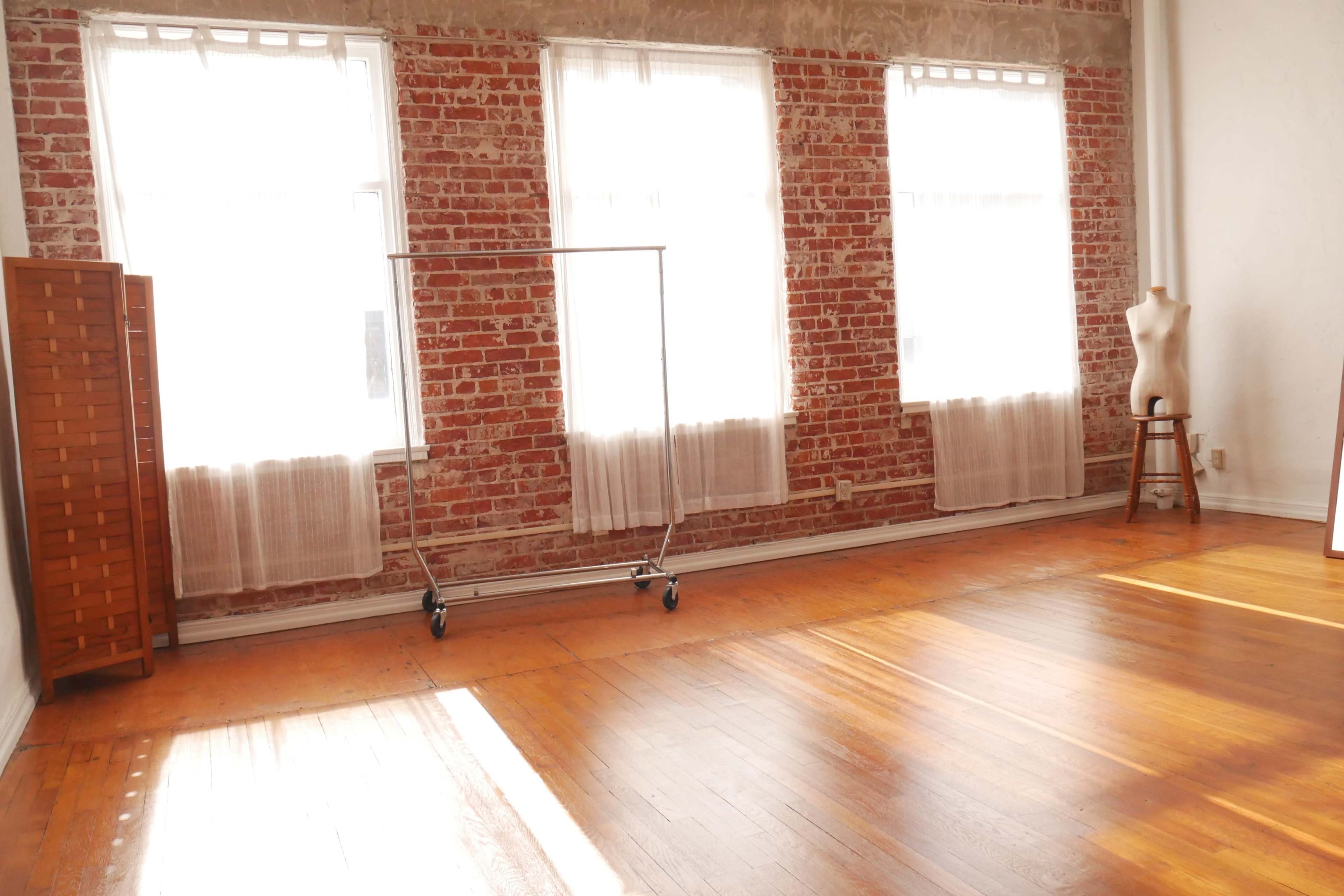 A bright, empty room with exposed brick walls, hardwood flooring, and large windows allowing natural light to pour in.