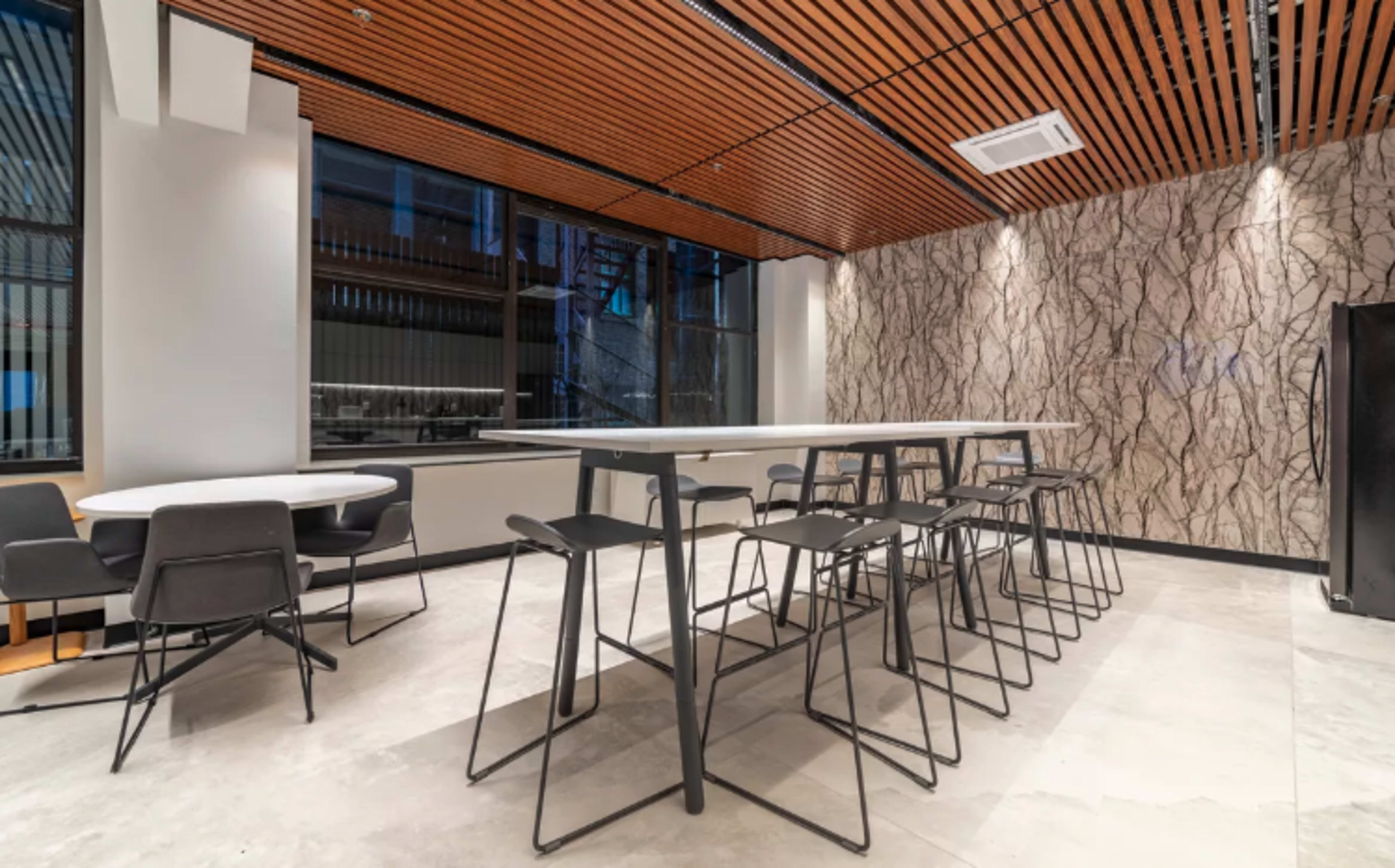 A modern dining area featuring long tables and black chairs, with a wood-paneled ceiling and textured wall.