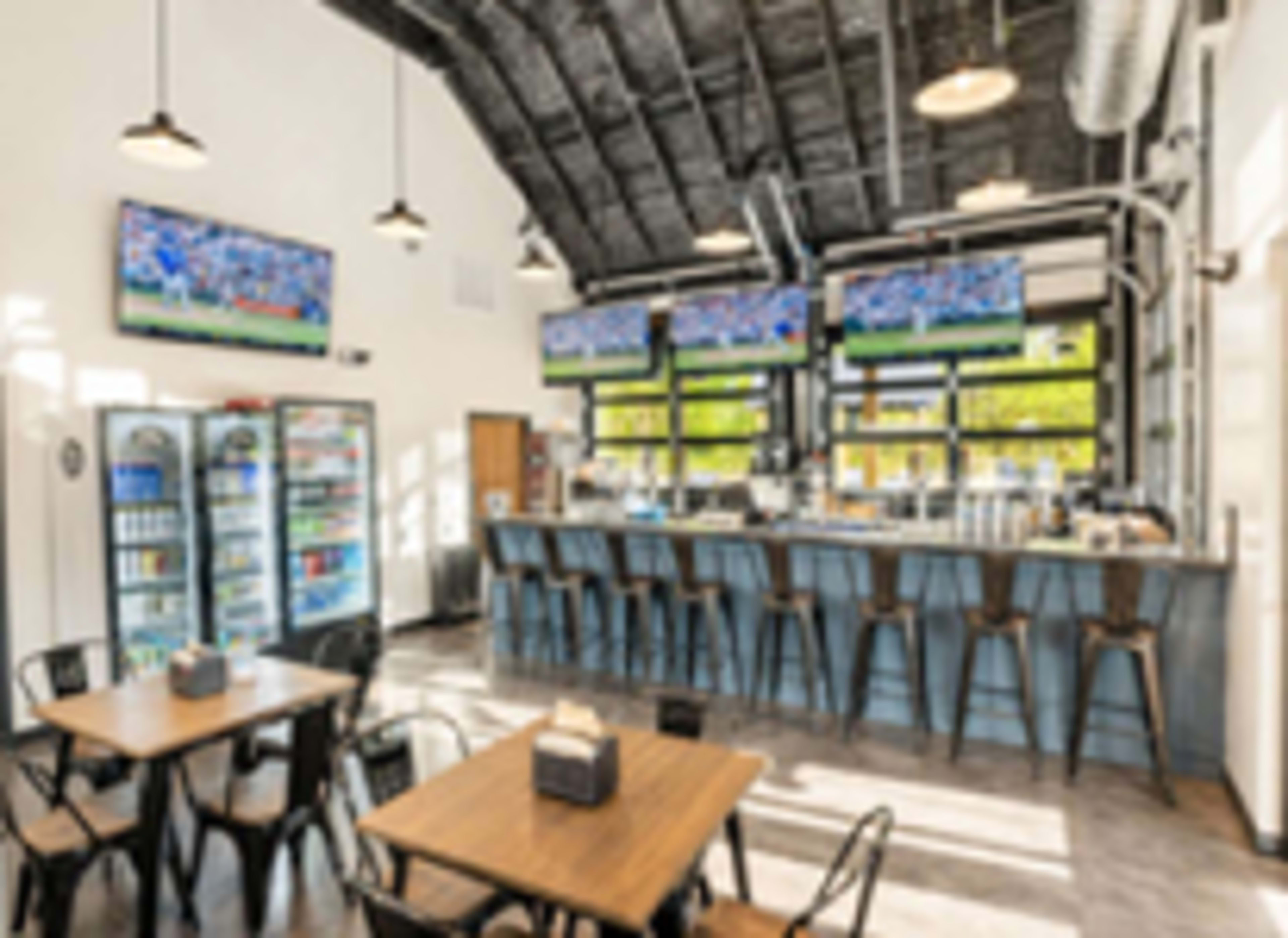 The image shows a modern bar and dining area with high ceilings, featuring several televisions mounted on the walls and a bar counter with stools, along with tables for seating.