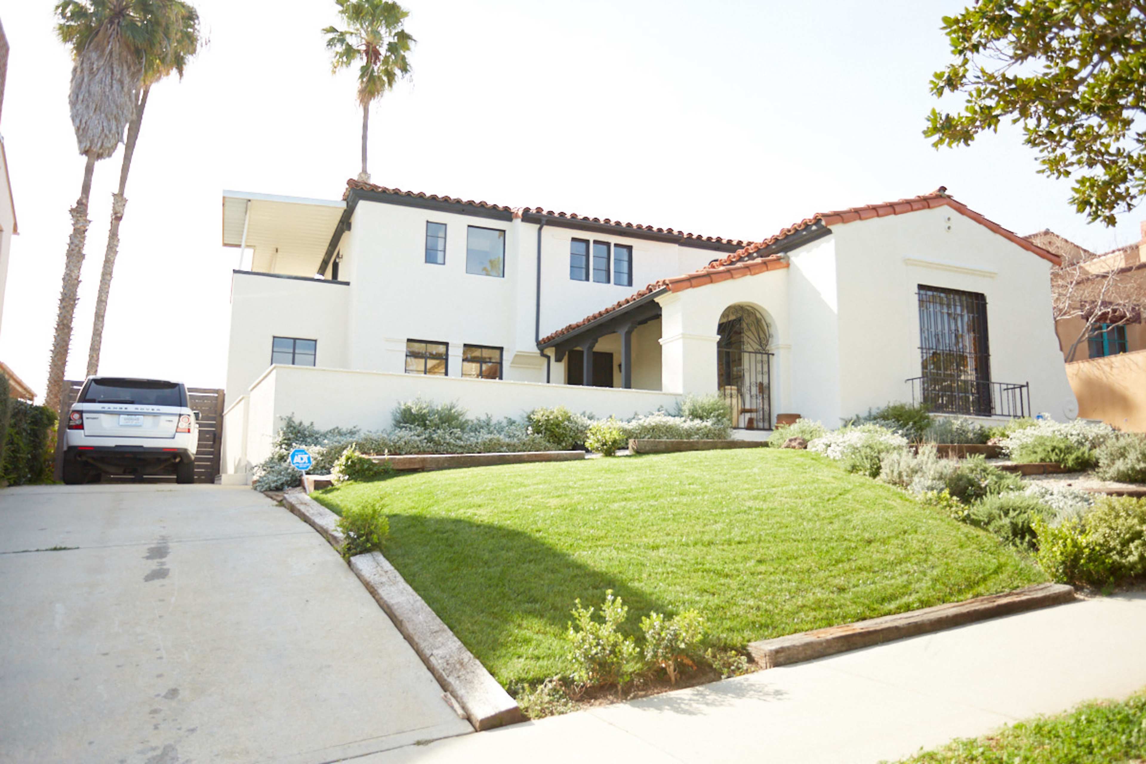 A white house with a red tile roof and a decorative archway sits on a landscaped lot with a sloped driveway and palm trees nearby.