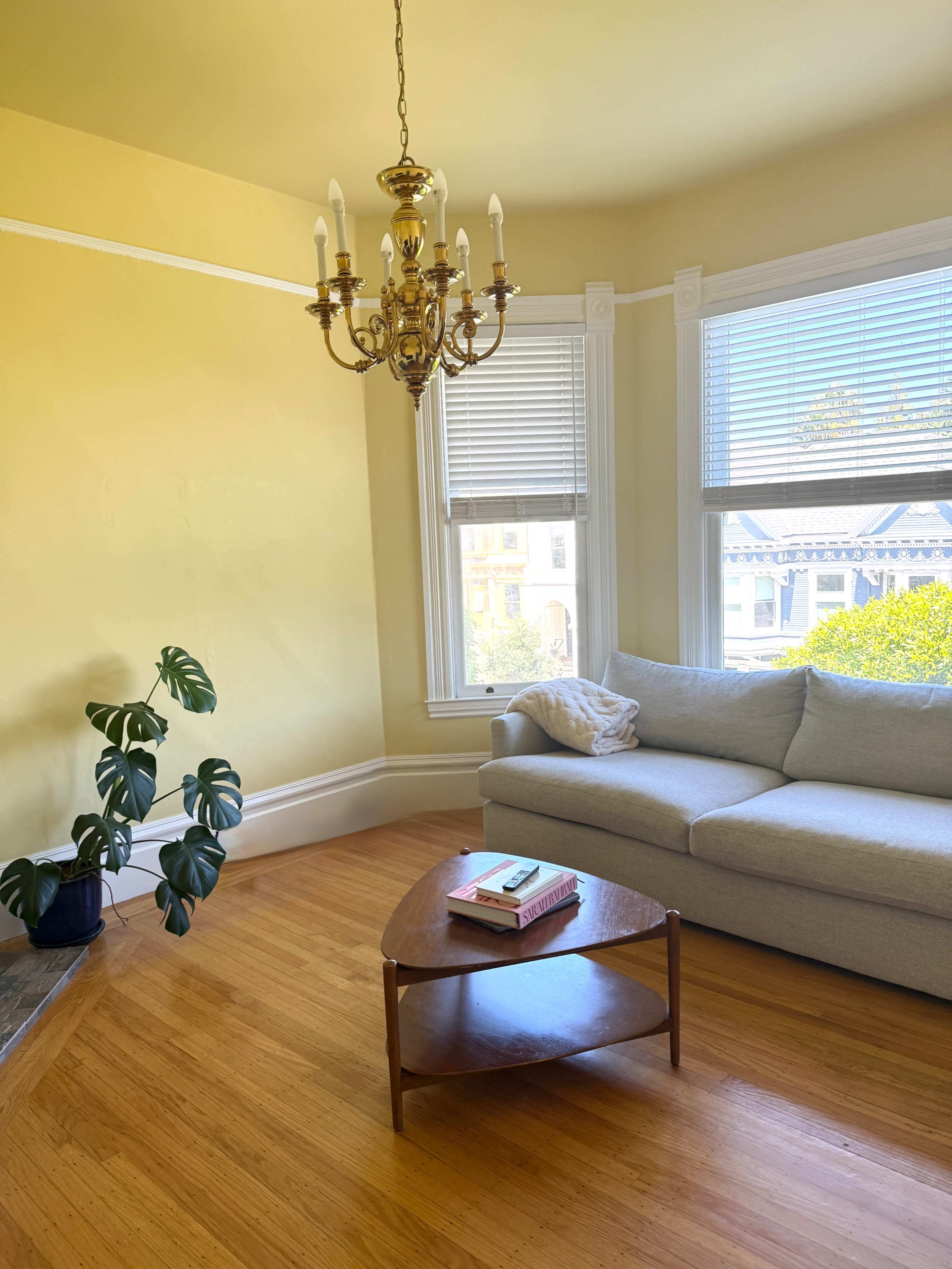 The image shows a bright living room with yellow walls, a chandelier, a gray sofa, a wooden coffee table, and a potted plant.