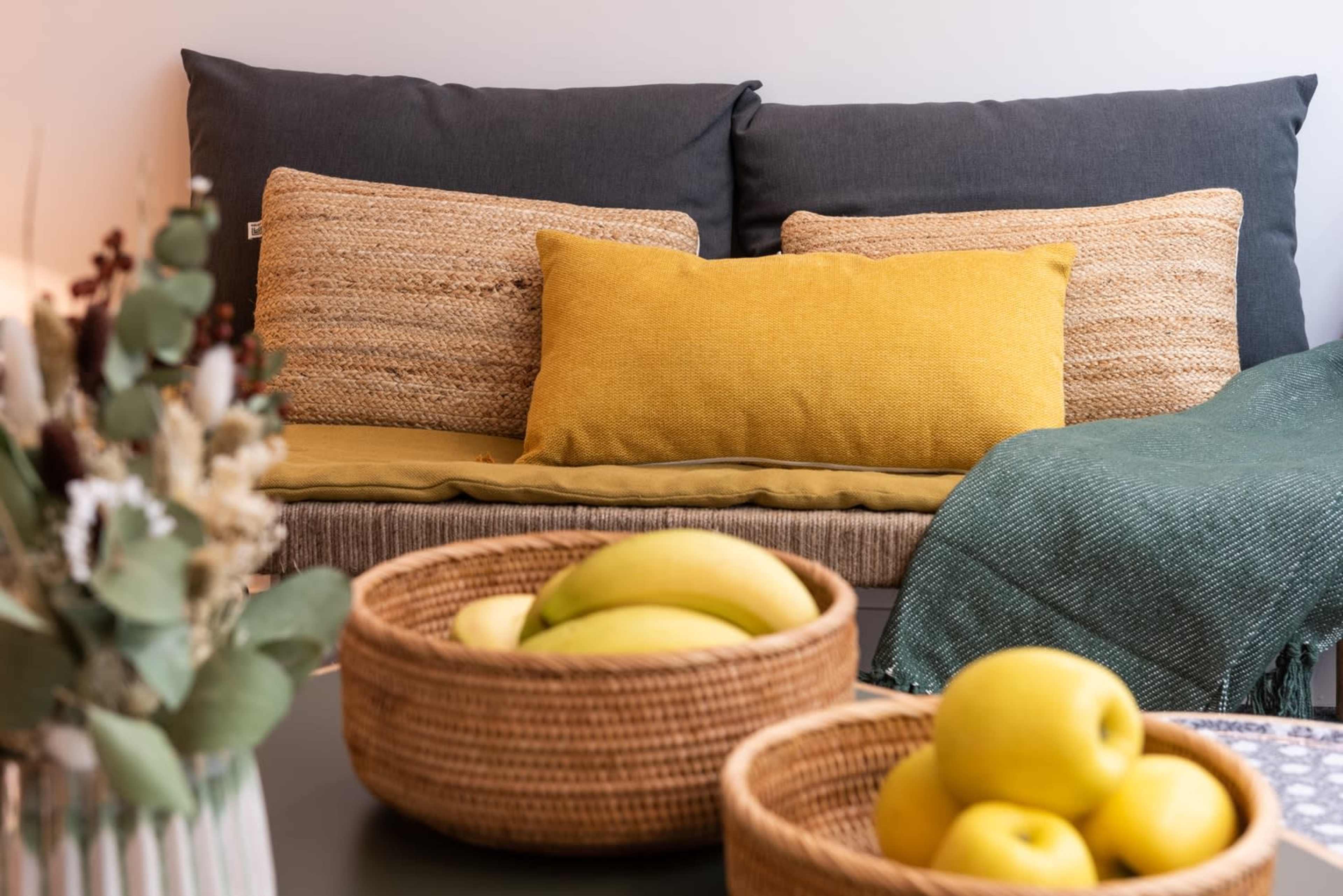 The image shows a cozy living room setup featuring a sofa with various cushions and two decorative woven baskets containing bananas and apples on a table.