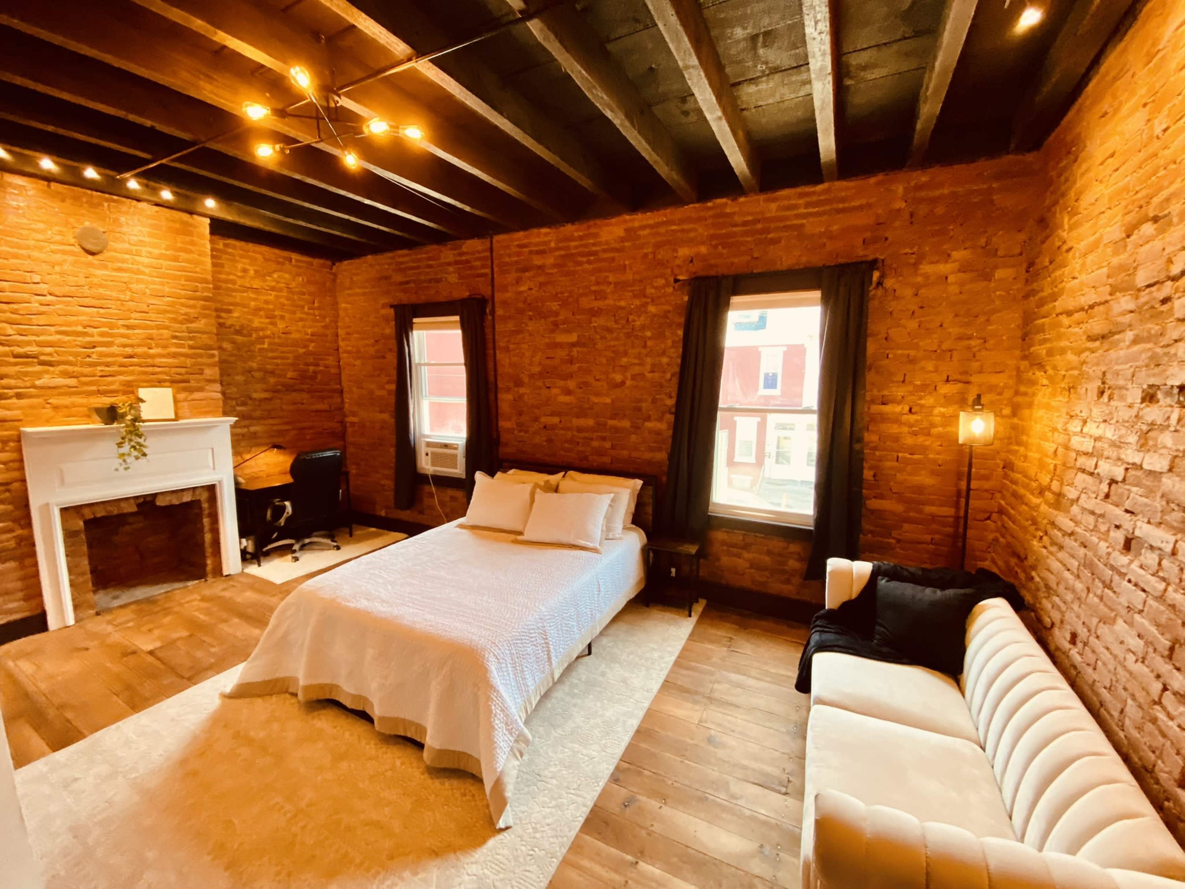A rustic bedroom with exposed brick walls, a bed, a white sofa, a small desk, and large windows allowing natural light.