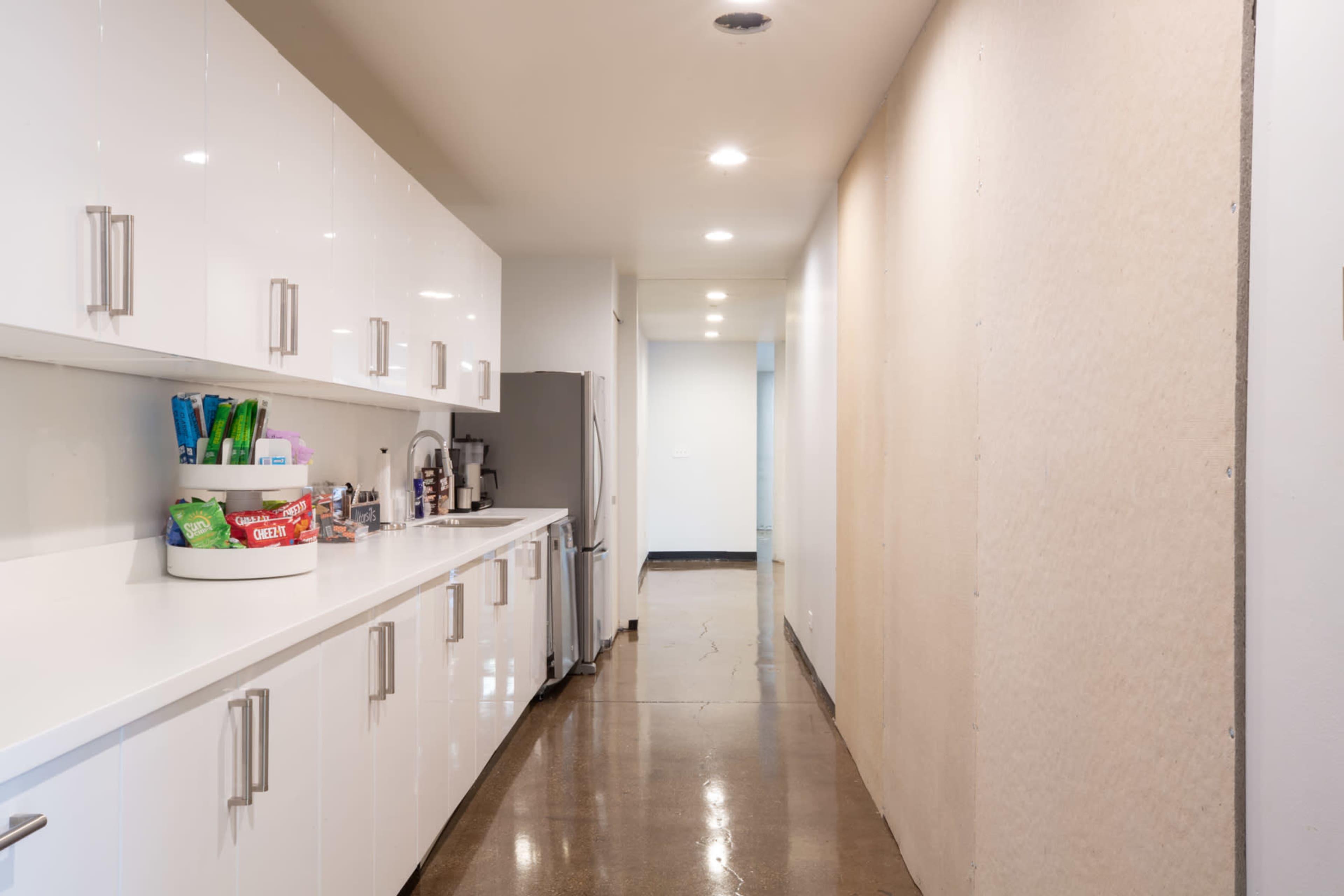 The image shows a wide hallway with glossy white cabinets on one side and a bare, unfinished wall on the other, featuring a polished floor.