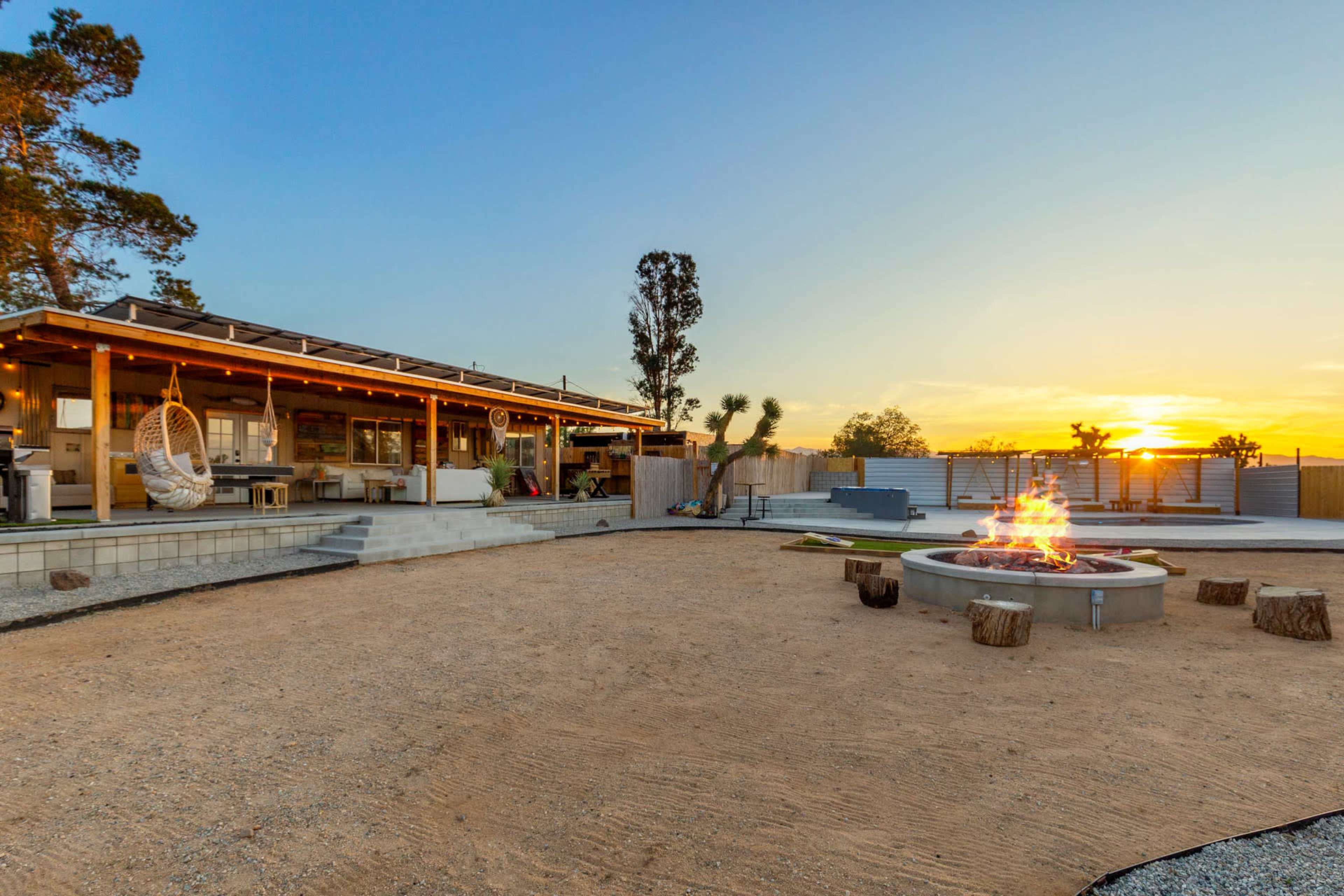 A modern outdoor space features a fire pit at the center, surrounded by seating, with a large building and sunset in the background.