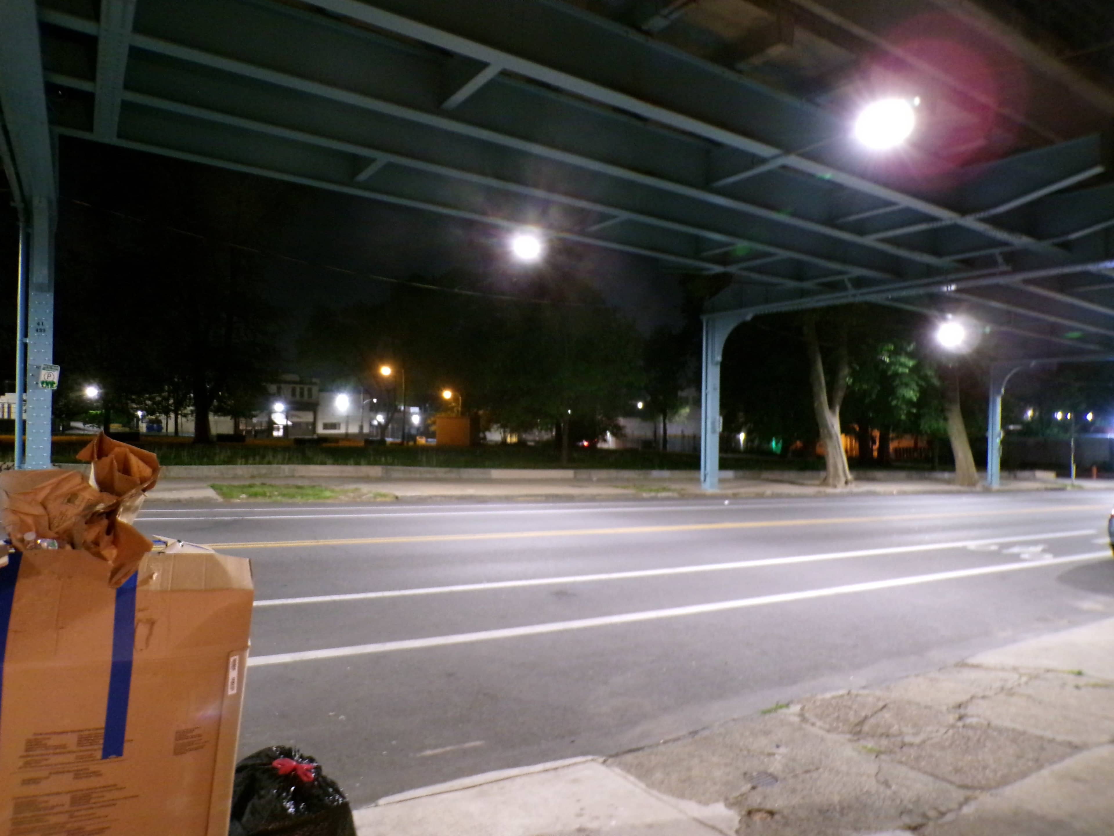 The image shows a dimly lit urban street beneath an elevated structure, with cardboard boxes and a trash bag visible in the foreground.