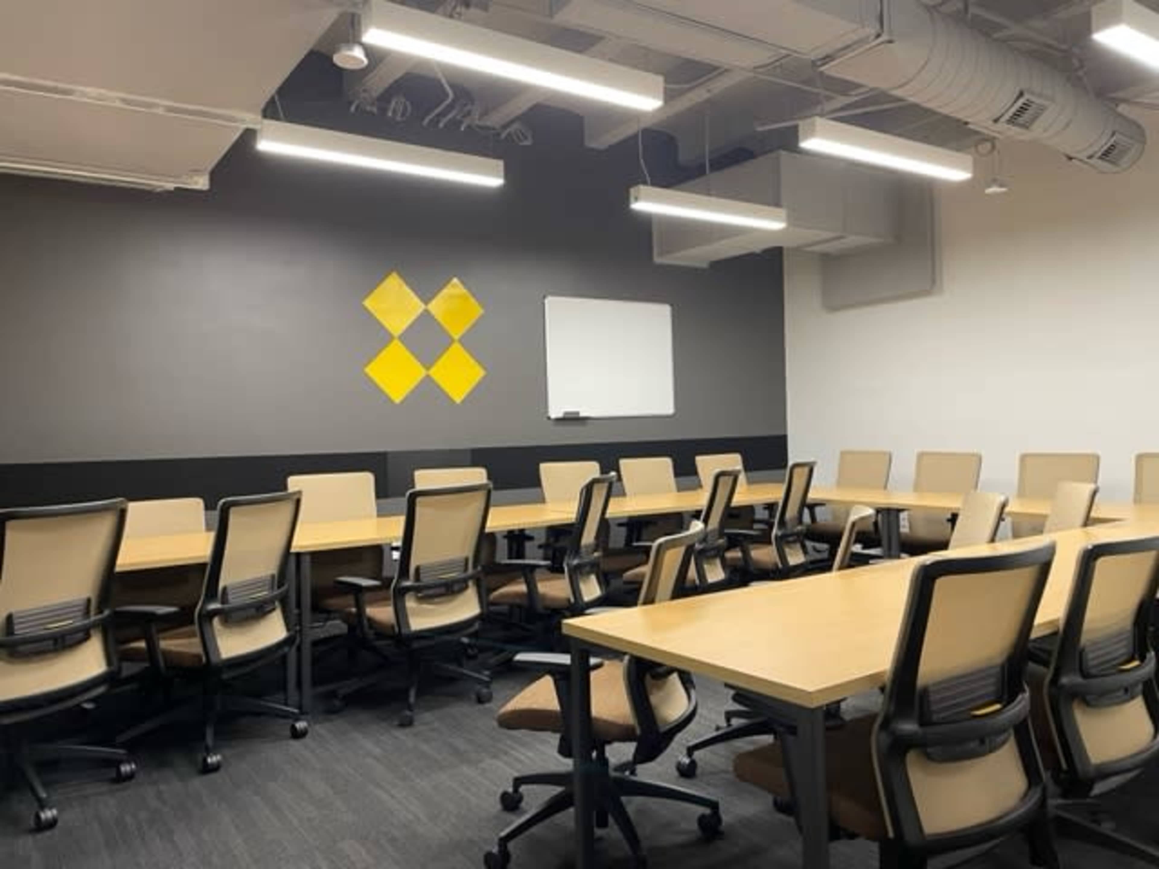 A conference room features multiple wooden tables and chairs arranged in rows, with a whiteboard and a yellow diamond graphic on the wall.